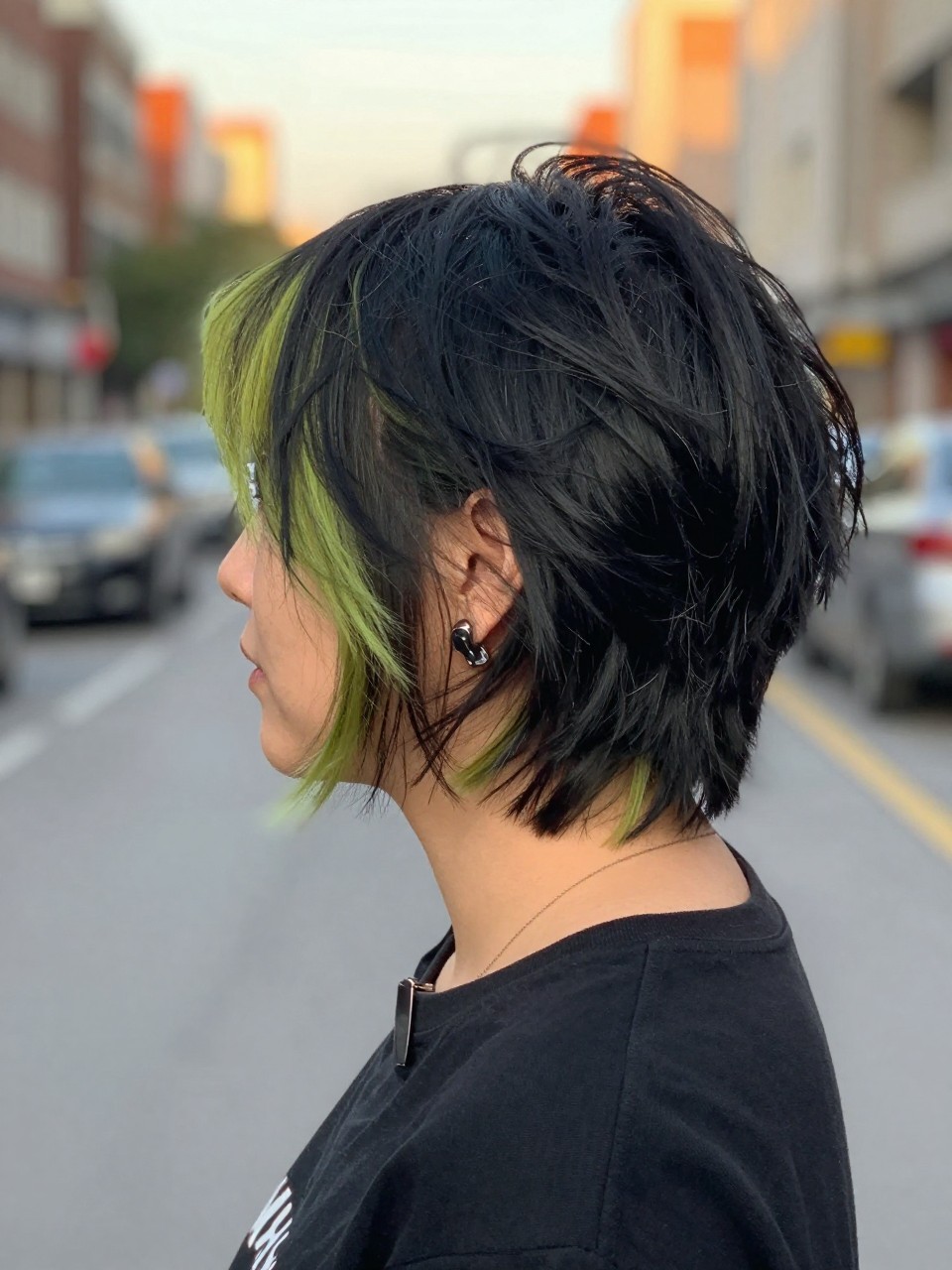 Photo of a woman wearing a tousled black bob with lime green highlights and soft choppy texture, back-of-head view to highlight details, in a softly blurred city street background, golden hour glow, containing a small lived-in detail such as a hair clip clipped to the collar and statement earrings peeking through, latest iPhone photo quality.