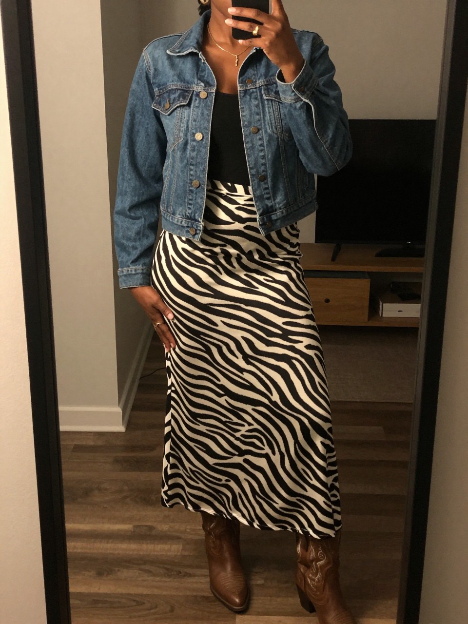mirror selfie of a black woman in a denim jacket and a zebra print long skirt with cowboy boots, one hand resting on her hip, cropped at NECK so face doesn't show, in a city apartment, warm moody lighting, iPhone photo quality.