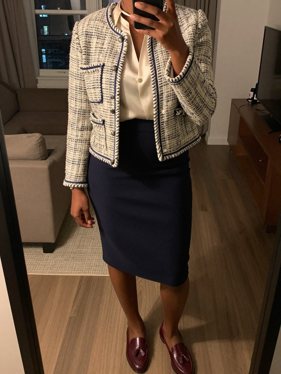 iPhone mirror selfie of a black woman wearing a cream and navy tweed jacket over a cream silk shell tucked into a navy pencil skirt with burgundy tassel loafers, one hand resting on her jacket, cropped at NECK so face doesn't show, in a city apartment, warm moody lighting, iPhone photo quality.