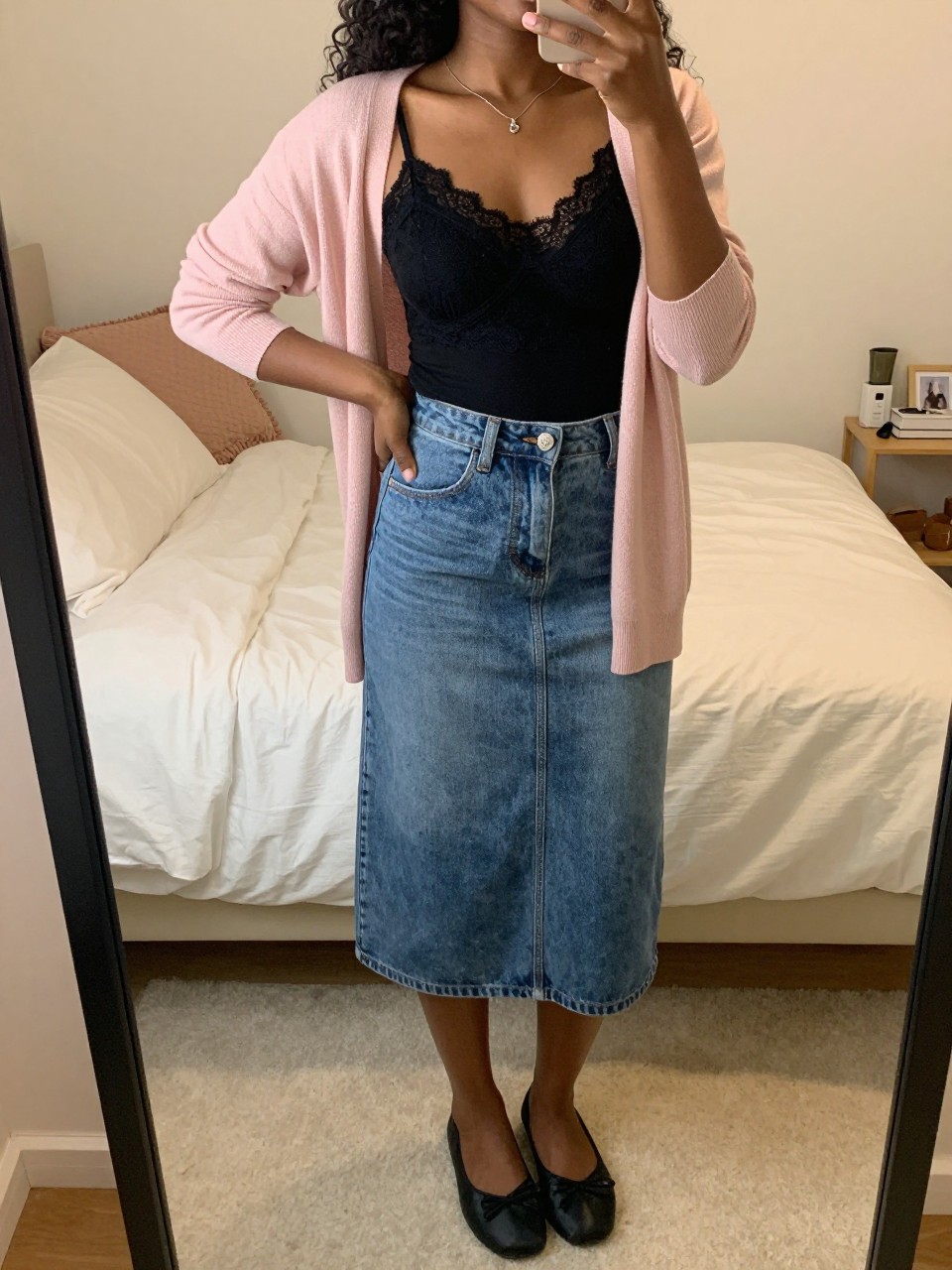 iPhone mirror selfie of a black woman wearing a black lace-trim camisole tucked into a mid-wash denim A-line skirt with a soft pink cardigan worn open and black ballet flats, standing in a sweet pose, cropped at NECK so face doesn't show, in a feminine-themed bedroom, warm moody lighting, iPhone photo quality.