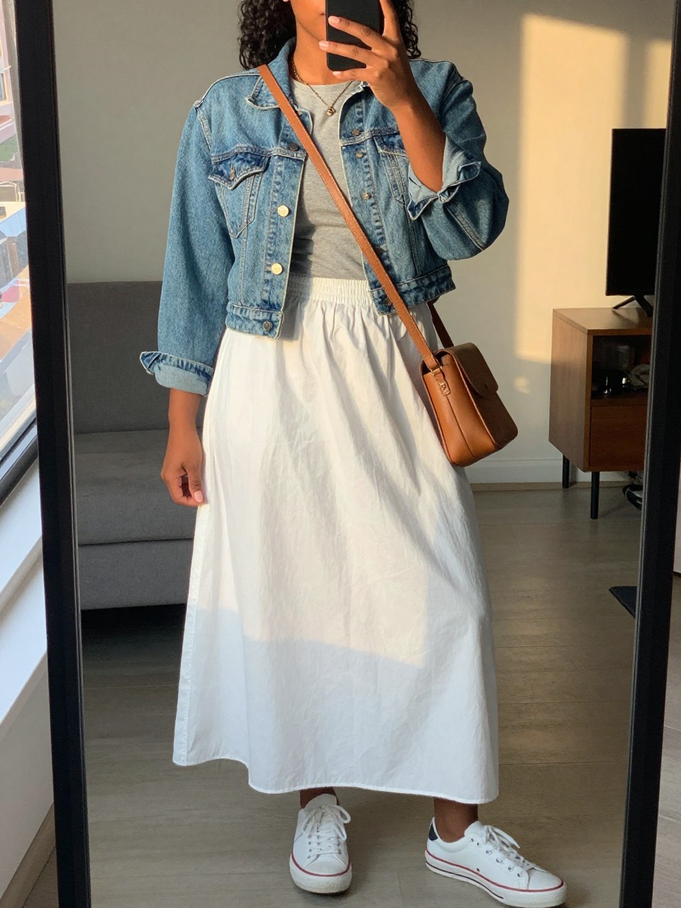 mirror selfie of a curvy black woman wearing a cropped light-wash denim jacket over a gray tee tucked into a white cotton maxi skirt with white sneakers and a tan leather crossbody bag, standing in a casual relaxed pose, cropped at NECK so face doesn't show, in a city apartment, evening golden hour, iPhone photo quality.