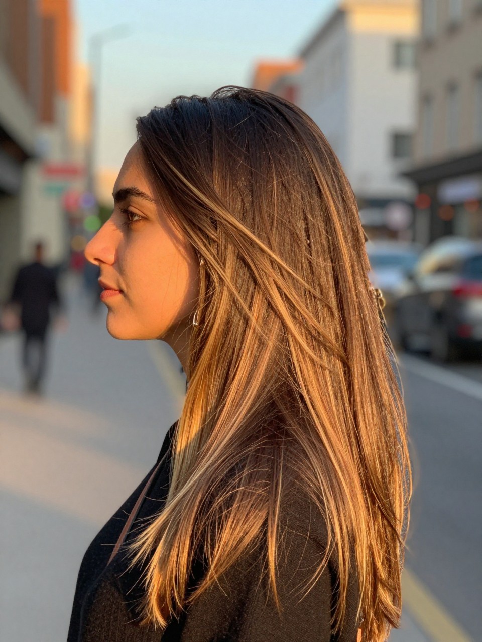 Photo of a woman wearing long straight brunette hair with blonde face-framing highlights, side profile view, in a softly blurred city street background, golden hour glow, containing a small lived-in detail such as statement earrings peeking through and a crossbody strap on the shoulder, latest iPhone photo quality.