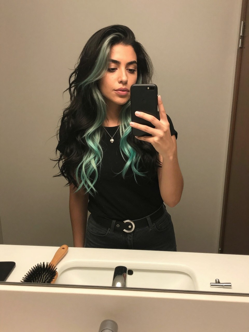 Photo of a mexican woman wearing a wavy black lob with mint green highlights, candid mirror selfie, at a bathroom vanity mirror, soft indoor lighting, containing a small lived-in detail such as a hairbrush on the sink and a silver clip nearby, latest iPhone photo quality.