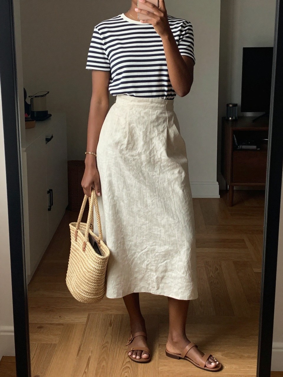 mirror selfie of a black woman wearing a navy striped Breton top tucked into a cream linen A-line midi skirt with flat tan sandals and a straw tote, one hand holding her phone at her side, cropped at NECK so face doesn't show, in a city apartment, warm moody lighting, iPhone photo quality.