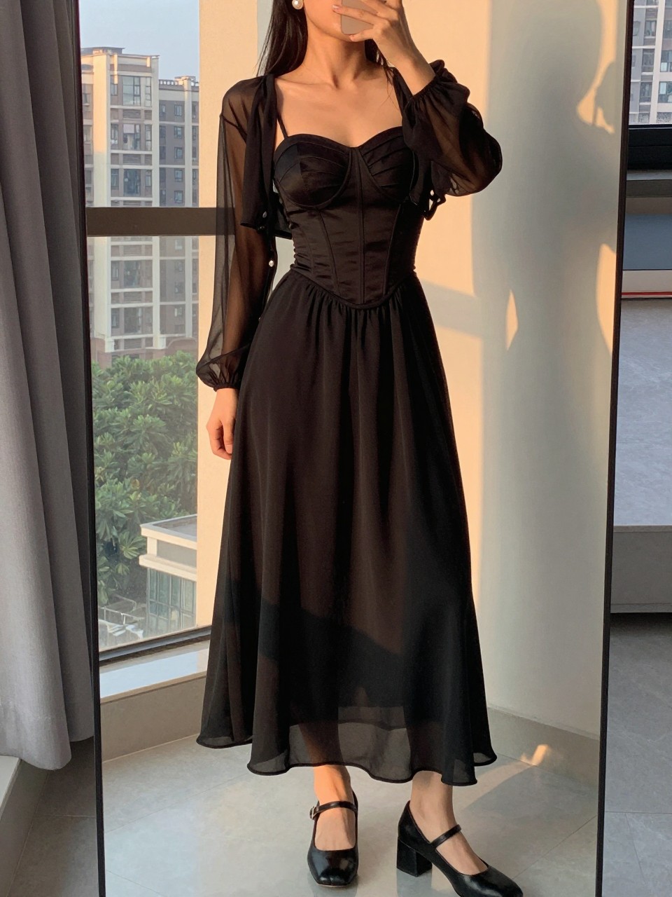 iPhone mirror selfie of a woman wearing a black corset top with a black chiffon maxi skirt, a sheer black mesh cardigan over top, and black heeled Mary Janes, pearl earrings visible, standing in a romantic edgy pose, cropped at NECK so face doesn't show, in a city apartment, evening golden hour, iPhone photo quality.