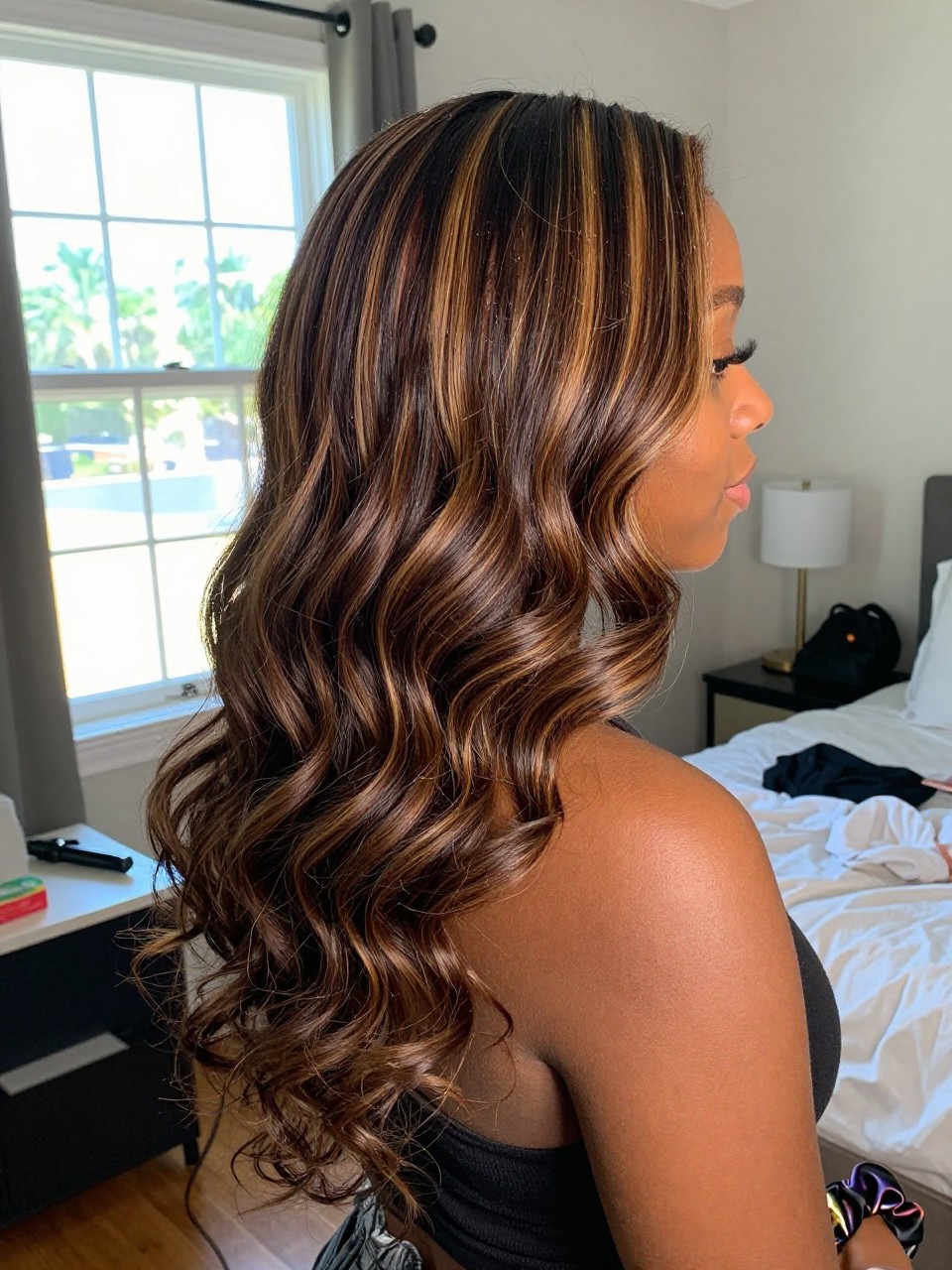 Photo of a Black woman wearing soft dark waves with honey brown highlight ribbons woven through the lengths, side profile view, in a sun-drenched bedroom, natural window light, containing a small lived-in detail such as a curling iron on the counter and a silk scrunchie on the wrist, latest iPhone photo quality.