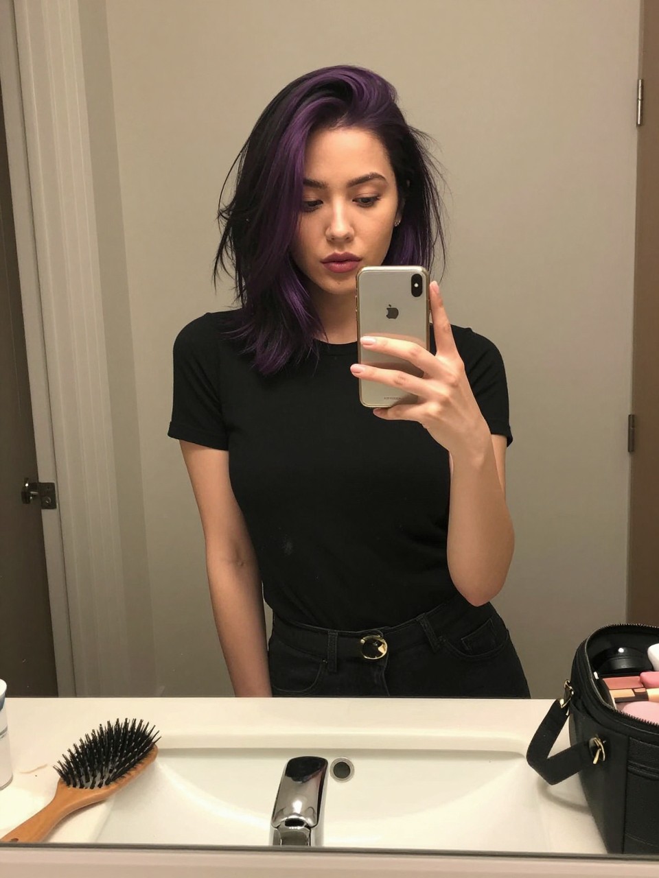 Photo of a woman wearing a sleek black lob with violet face-framing highlights, candid mirror selfie, at a bathroom vanity mirror, soft indoor lighting, containing a small lived-in detail such as a hairbrush on the vanity and a makeup bag nearby, latest iPhone photo quality.