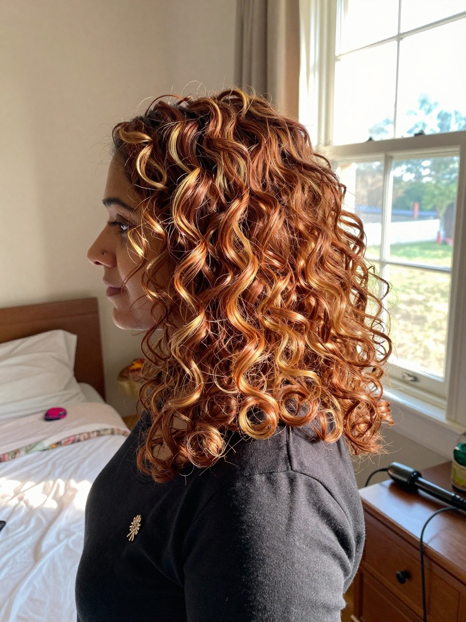 Photo of a indian woman wearing soft copper curls with golden blonde highlights, side profile view, in a sun-drenched bedroom, natural window light, containing a small lived-in detail such as a decorative pin on one side and a curling iron resting on the nightstand, latest iPhone photo quality.