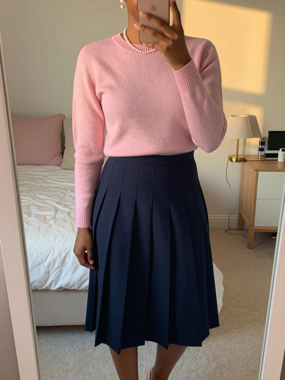 iPhone mirror selfie of a black woman wearing a baby pink crewneck sweater tucked into a navy pleated midi skirt with pearl earrings and nude heels, one hand gently touching her pearls, cropped at NECK so face doesn't show, in a feminine-themed bedroom, evening golden hour, iPhone photo quality.