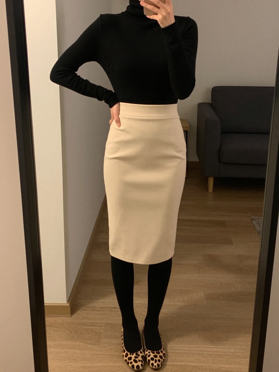mirror selfie of a woman wearing a black turtleneck tucked into a soft cream pencil skirt with leopard print flats and black tights, one hand on her hip, cropped at NECK so face doesn't show, in a city apartment, warm moody lighting, iPhone photo quality.
