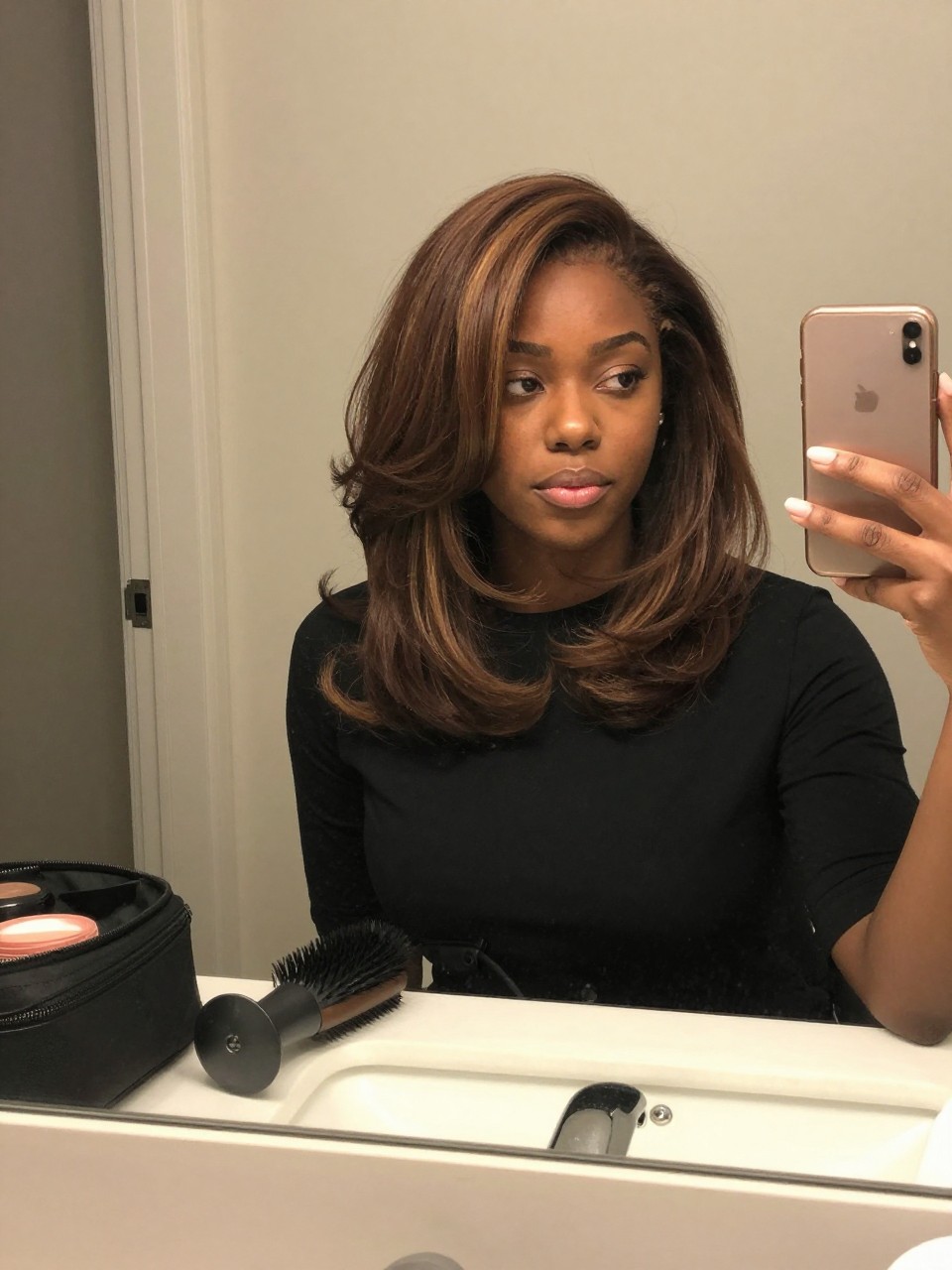 Photo of a black woman wearing chocolate brown hair with cinnamon lowlights in a soft layered blowout, candid mirror selfie, bathroom vanity mirror setting, soft indoor lighting, with a blow dryer brush on the counter and a makeup bag nearby, latest iPhone photo quality.