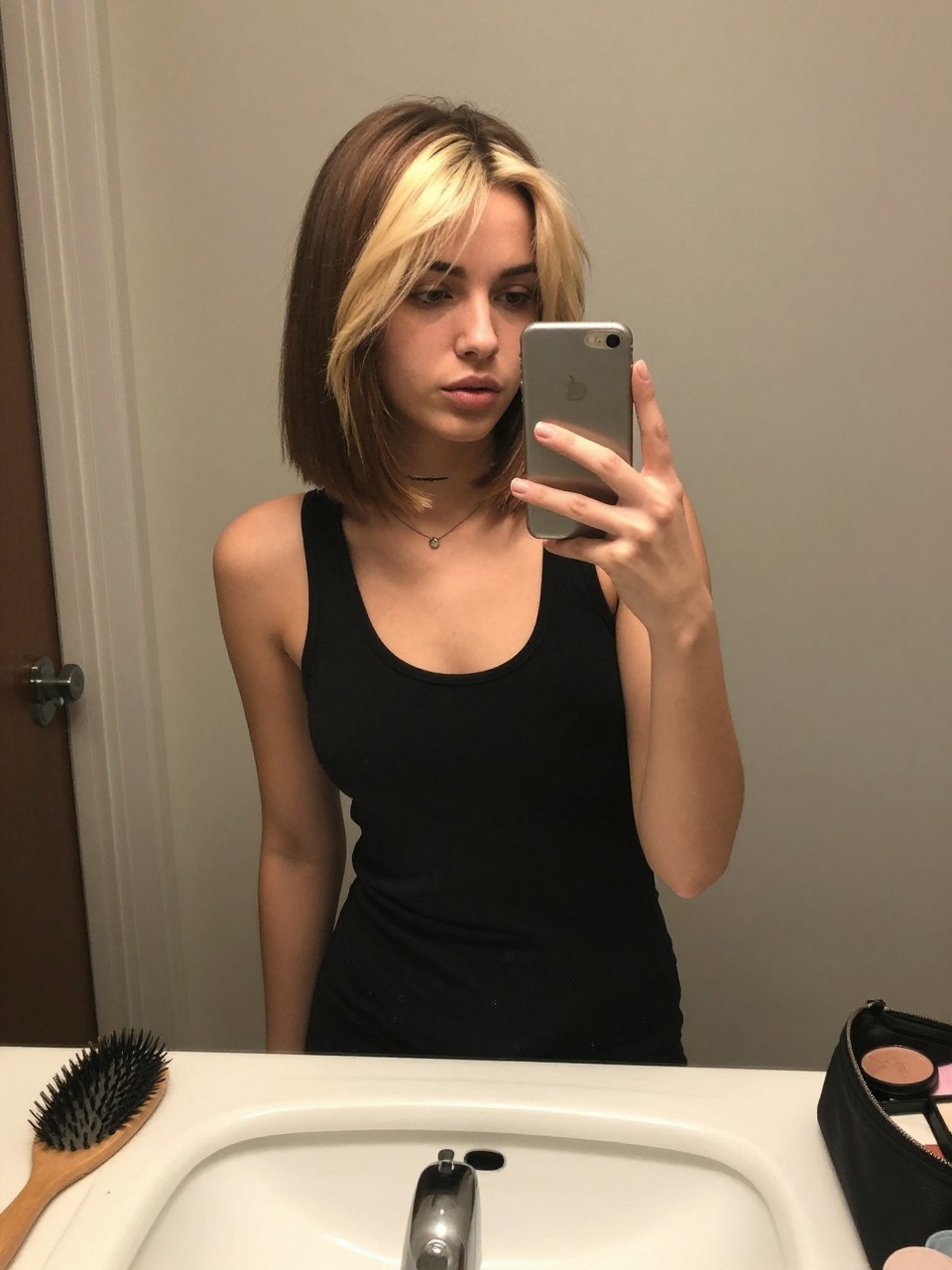 Photo of a BIRACIAL woman wearing a straight short brown bob with bold blonde face-framing highlights, candid mirror selfie, at a bathroom vanity mirror, soft indoor lighting, containing a small lived-in detail such as a hairbrush on the sink and a makeup bag nearby, latest iPhone photo quality.