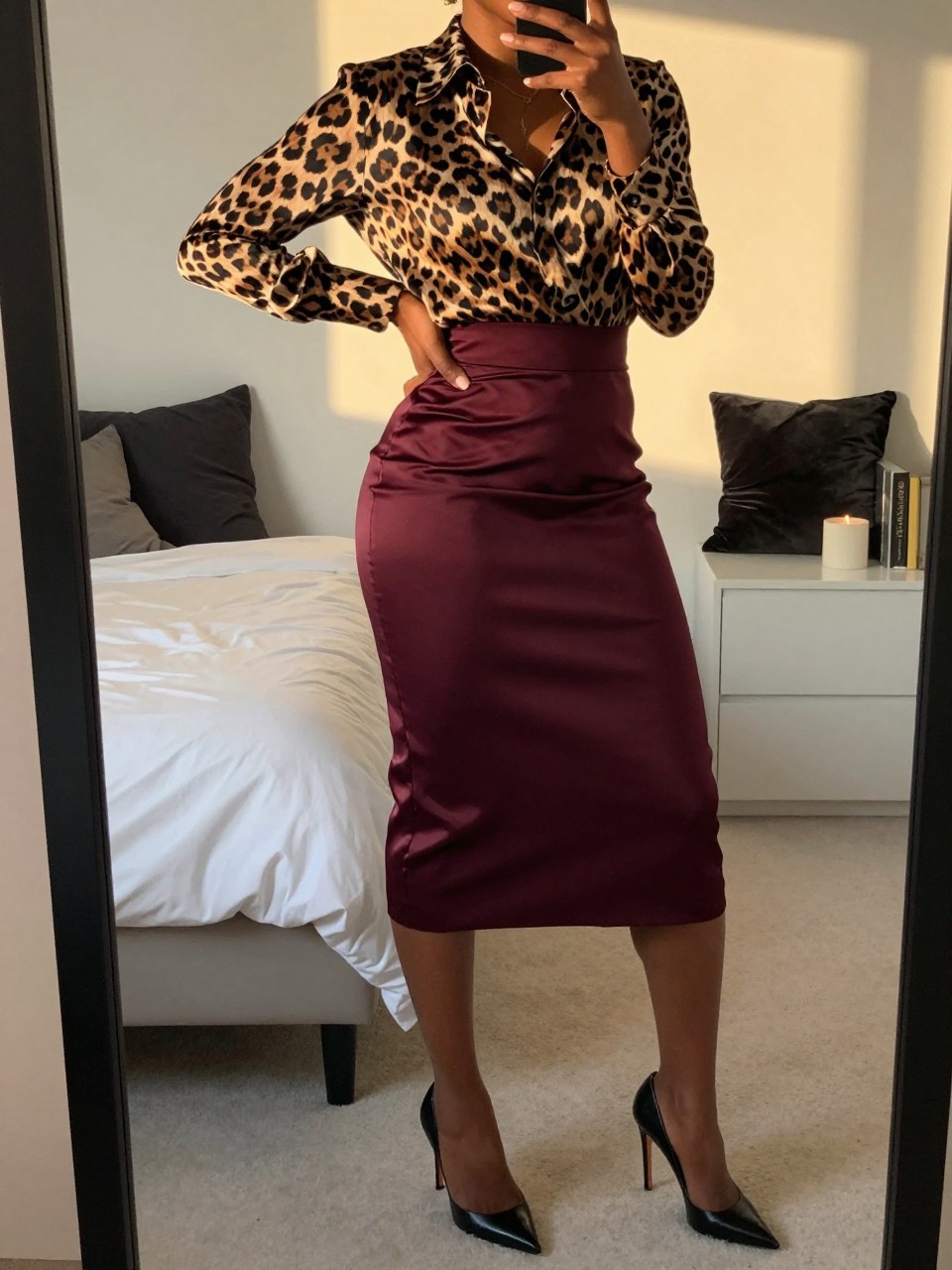 mirror selfie of a black woman wearing a leopard print silk blouse tucked into a deep burgundy satin pencil skirt with black stiletto heels, one hand on her hip, cropped at NECK so face doesn't show, in a feminine-themed bedroom, evening golden hour, iPhone photo quality.