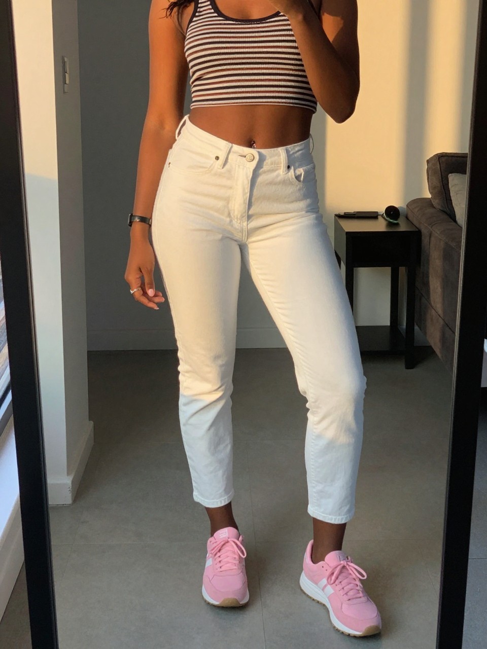 iPhone mirror selfie of a black woman in white jeans and a striped top with pink sneakers, standing with one foot slightly forward, cropped at NECK so face doesn't show, in a city apartment, evening golden hour, iPhone photo quality.