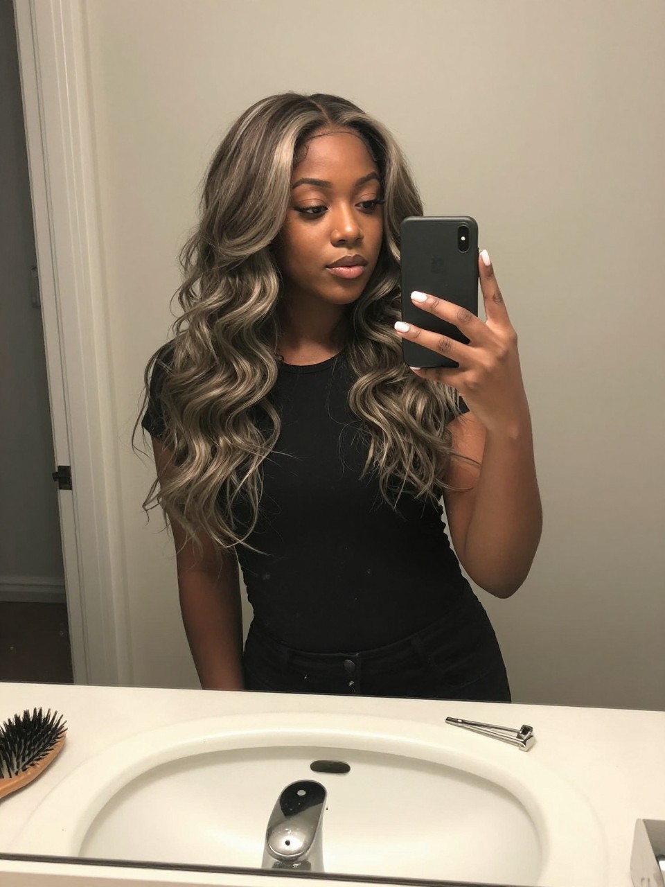 Photo of a black woman wearing a wavy ash brown lob with silver beige highlights, candid mirror selfie, at a bathroom vanity mirror, soft indoor lighting, containing a small lived-in detail such as a hairbrush on the sink and a silver clip nearby, latest iPhone photo quality.