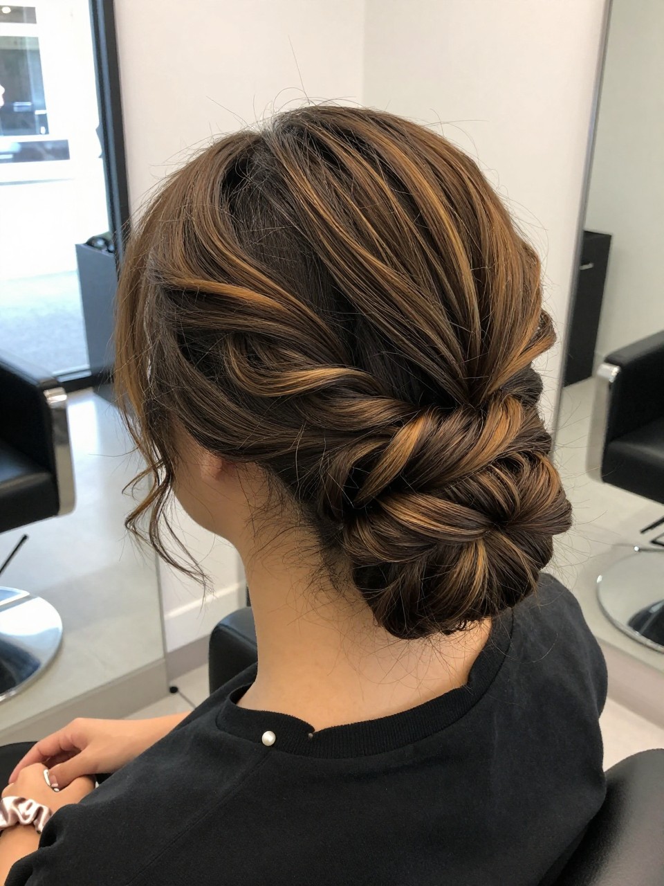 Photo of a back woman wearing a low textured bun on dark hair with honey highlights woven through the twists, back-of-head view to highlight details, in a hair salon , natural window light, containing a small lived-in detail such as a pearl pin and a silk scrunchie on the wrist, latest iPhone photo quality.