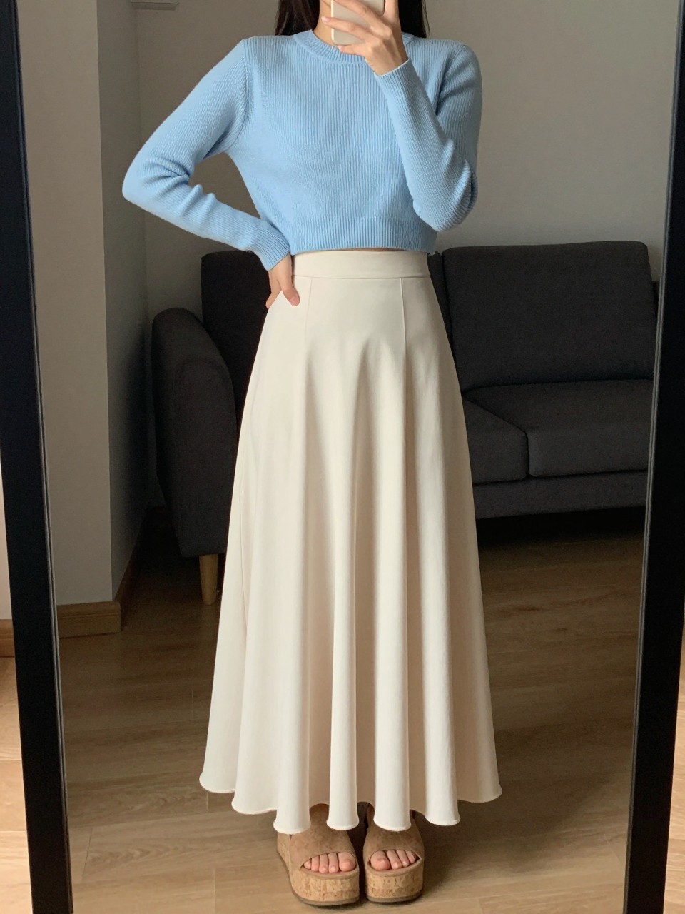 iPhone mirror selfie of a woman wearing a baby blue cropped ribbed sweater with a high-waisted soft cream flowy maxi skirt and cork platform sandals, one hand on her hip, cropped at NECK so face doesn't show, in a city apartment, warm moody lighting, iPhone photo quality.