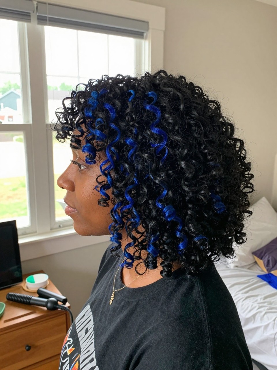 Photo of a black woman wearing soft black curls with sapphire blue highlight streaks, side profile view, in a sun-drenched bedroom, natural window light, containing a small lived-in detail such as a decorative pin on one side and a curling iron resting on the nightstand, latest iPhone photo quality.