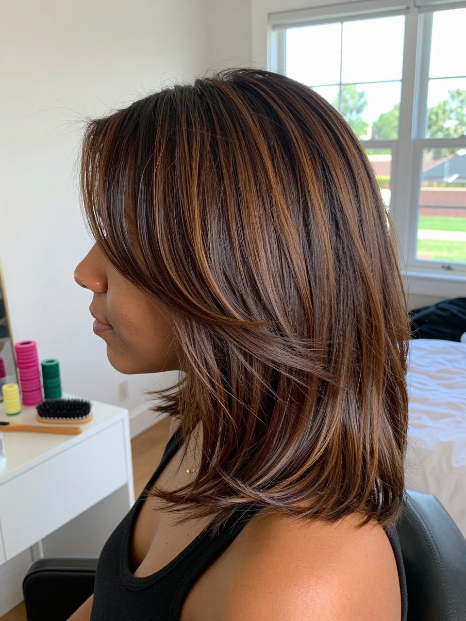 Photo of a biracial woman wearing a side part brown blowout with deep cinnamon dark highlights, side profile view, in a sun-drenched bedroom, natural window light, containing a small lived-in detail such as velcro rollers on the vanity and a round brush nearby, latest iPhone photo quality.