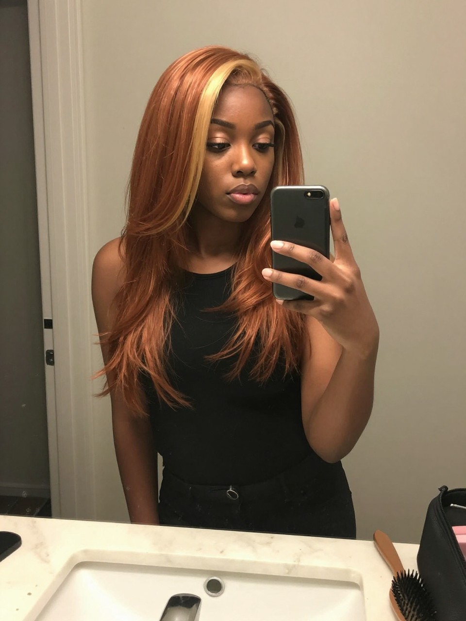 Photo of a black woman wearing a sleek copper lob with blonde face-framing highlights, candid mirror selfie, at a bathroom vanity mirror, soft indoor lighting, containing a small lived-in detail such as a hairbrush on the vanity and a makeup bag nearby, latest iPhone photo quality.