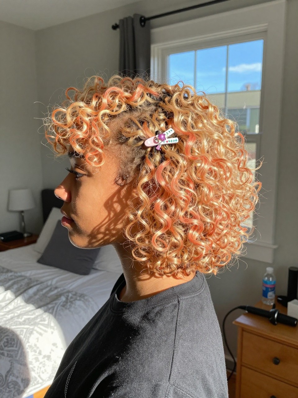 Photo of a biracial woman wearing side part strawberry blonde curls with apricot highlights, side profile view, in a sun-drenched bedroom, natural window light, containing a small lived-in detail such as a decorative clip on one side and a curling iron resting on the nightstand, latest iPhone photo quality.