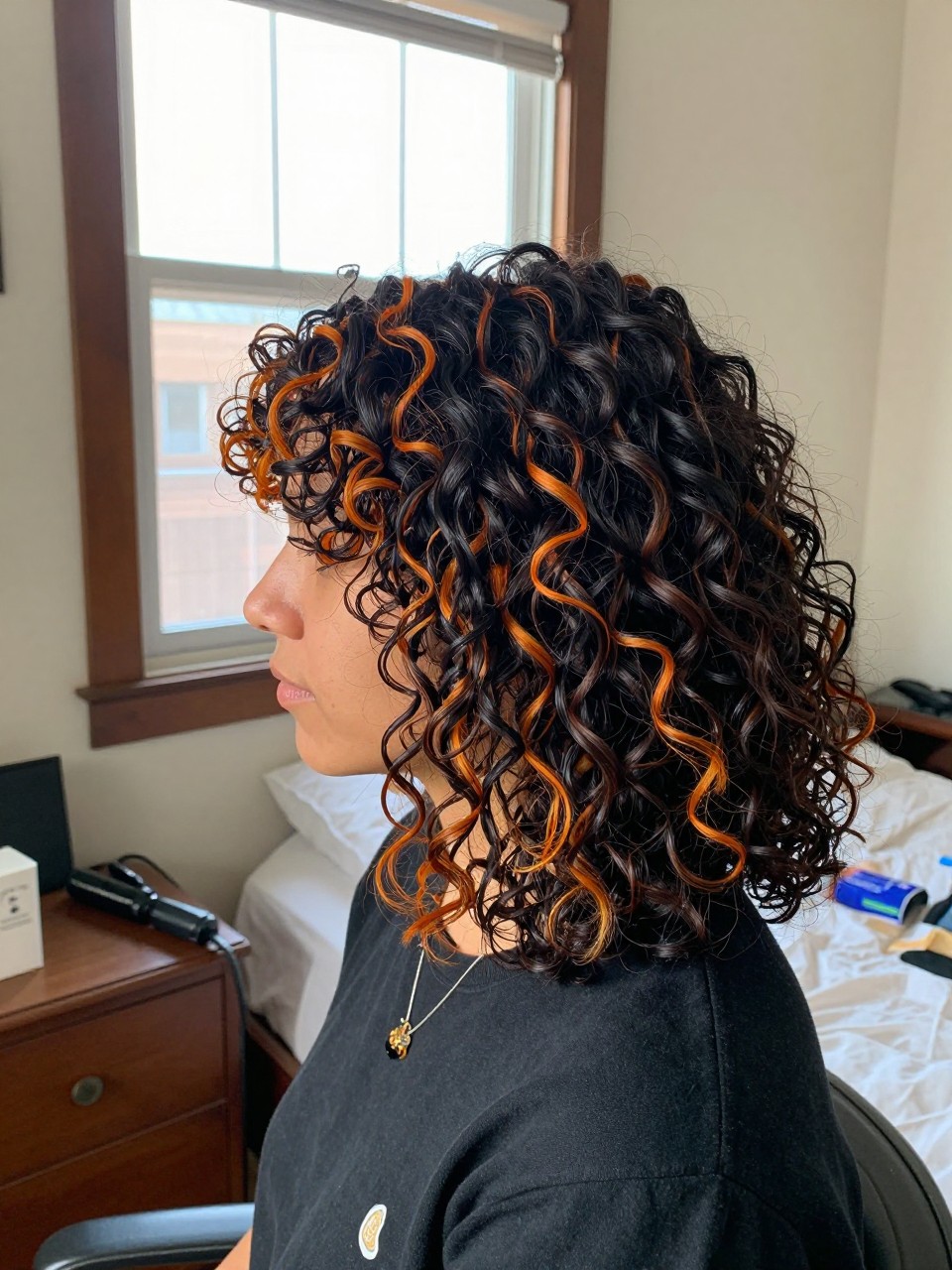 Photo of a woman wearing soft black curls with pumpkin orange highlight streaks, side profile view, in a sun-drenched bedroom, natural window light, containing a small lived-in detail such as a decorative pin on one side and a curling iron resting on the nightstand, latest iPhone photo quality.