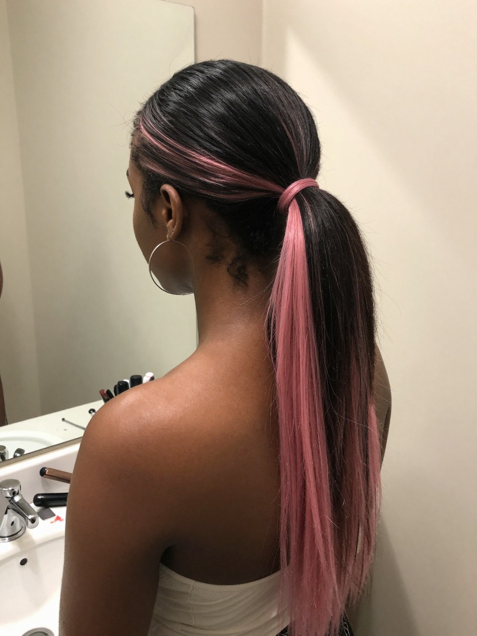 Photo of a black woman wearing a sleek mid ponytail with black hair and pink highlight panels through the tail, back-of-head view to highlight details, at a bathroom vanity mirror, soft indoor lighting, containing a small lived-in detail such as a flat iron on the counter and hoop earrings peeking through, latest iPhone photo quality.