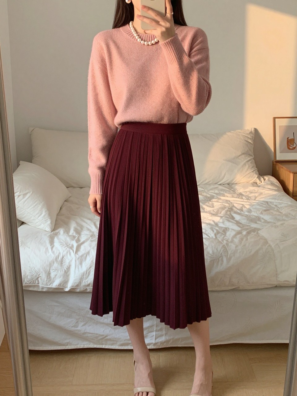 iPhone mirror selfie of a woman wearing a blush pink crewneck sweater tucked into a deep burgundy pleated midi skirt with pearl earrings and nude block-heel sandals, one hand gently touching her pearls, cropped at NECK so face doesn't show, in a feminine-themed bedroom, evening golden hour, iPhone photo quality.