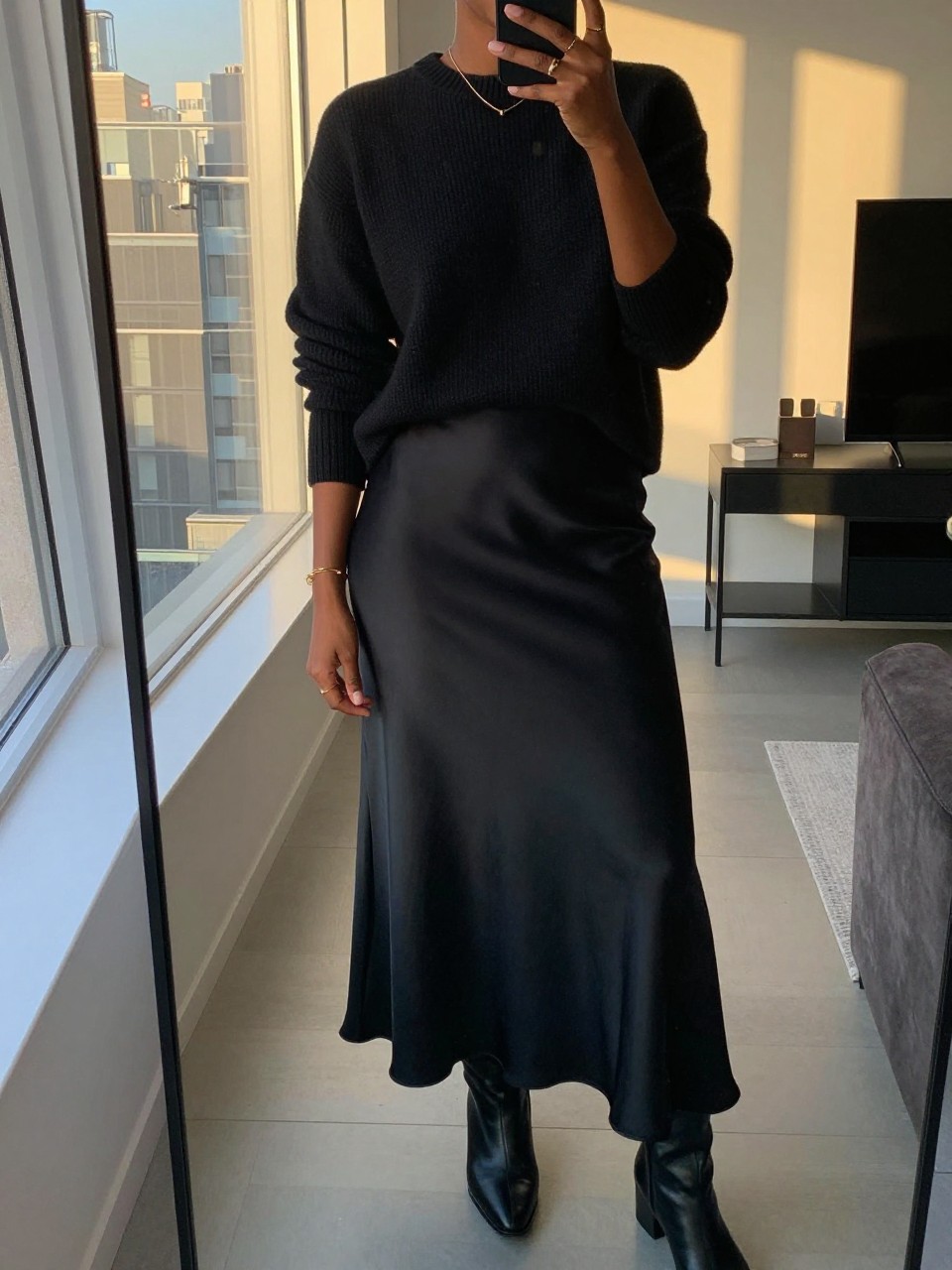 iPhone mirror selfie of a black woman wearing a black cashmere crewneck sweater tucked into a black satin maxi skirt creating a monochrome look, with black leather ankle boots and minimal gold jewelry, standing in a sophisticated pose, cropped at NECK so face doesn't show, in a city apartment, evening golden hour, iPhone photo quality.