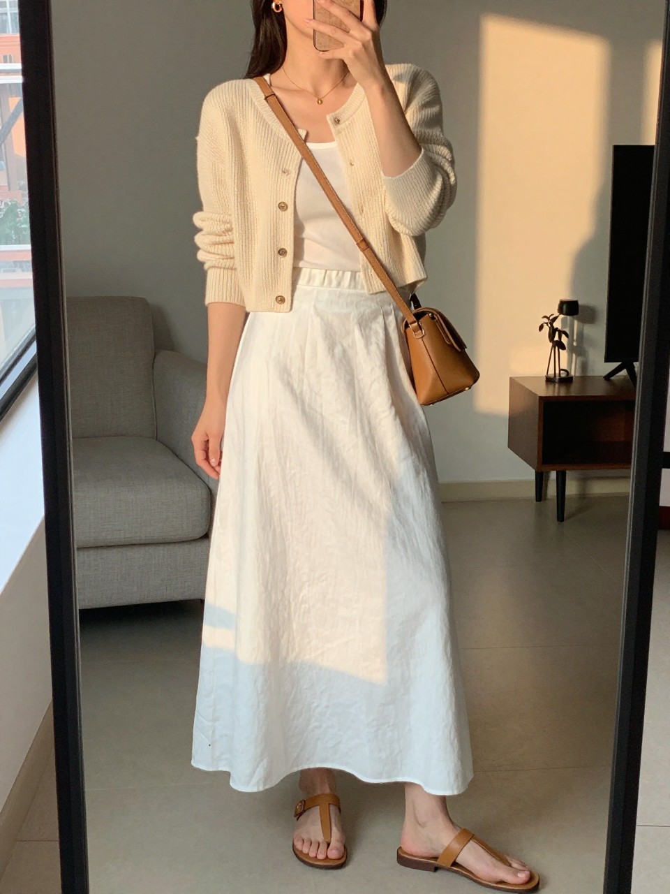 mirror selfie of a woman wearing a cream cropped cardigan over a white tank top tucked into a white linen maxi skirt with flat tan leather sandals and small gold hoop earrings, holding a tan crossbody bag, standing in a relaxed elegant pose, cropped at NECK so face doesn't show, in a city apartment, evening golden hour, iPhone photo quality.