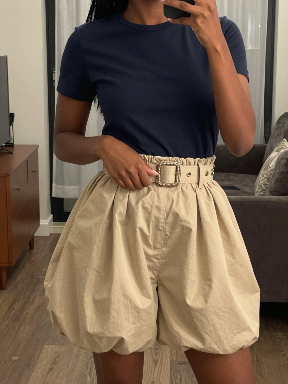 mirror selfie of a black woman in a navy t-shirt and beige balloon skirt with a statement belt, hand resting on the belt buckle, cropped at NECK so face doesn't show, in a city apartment, warm moody lighting, iPhone photo quality.
