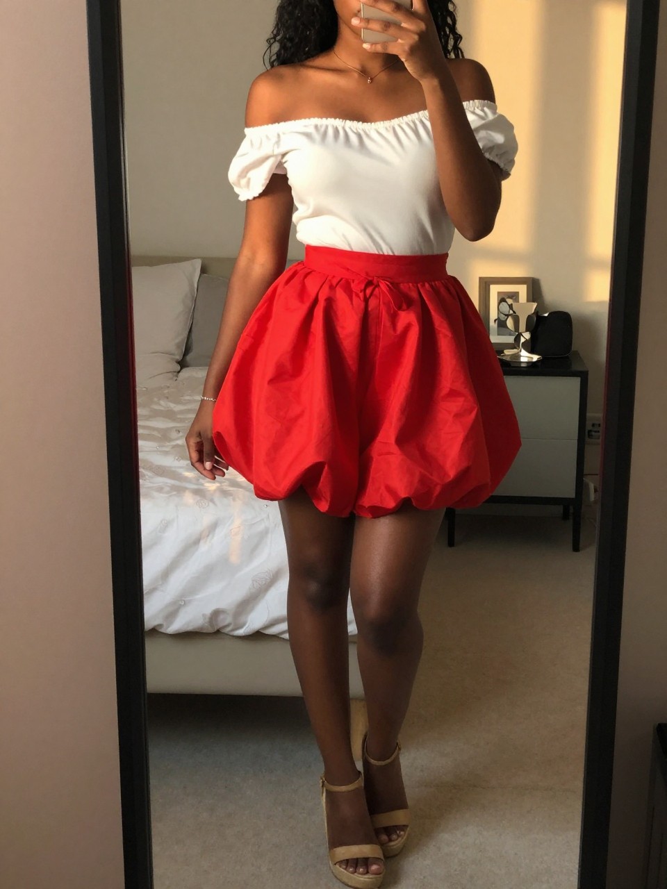 mirror selfie of a black woman in an off-the-shoulder top and a red balloon skirt with wedge sandals, posing with one hand gently touching her hair, cropped at NECK so face doesn't show, in a feminine-themed bedroom, evening golden hour, iPhone photo quality.