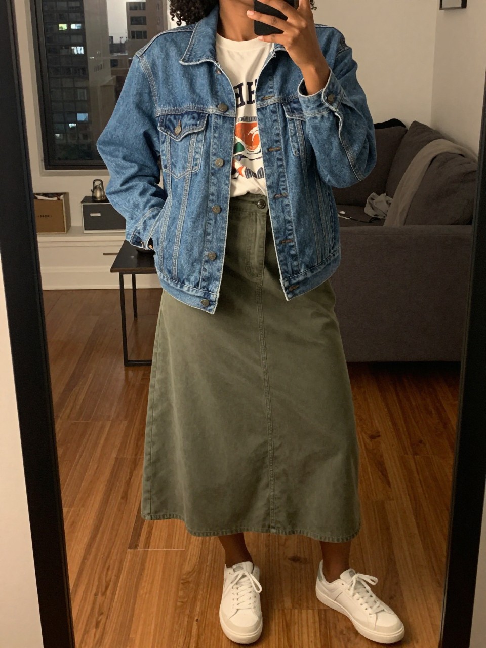 mirror selfie of a black woman wearing a blue denim jacket over a graphic tee with an olive green cotton midi skirt and white sneakers, one hand in her jacket pocket, cropped at NECK so face doesn't show, in a city apartment, warm moody lighting, iPhone photo quality.