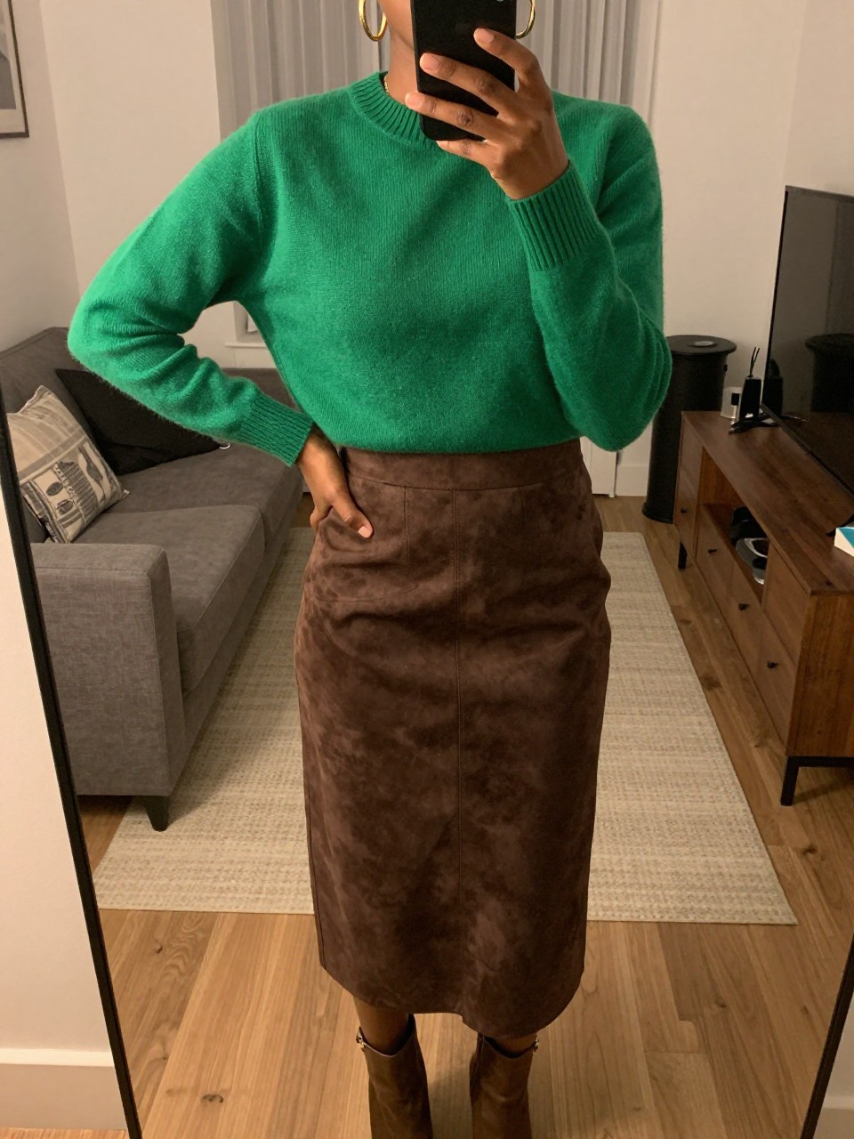 iPhone mirror selfie of a black woman wearing an emerald green crewneck sweater tucked into a chocolate brown suede midi skirt with gold hoop earrings and brown ankle boots, one hand on her hip, cropped at NECK so face doesn't show, in a city apartment, warm moody lighting, iPhone photo quality.