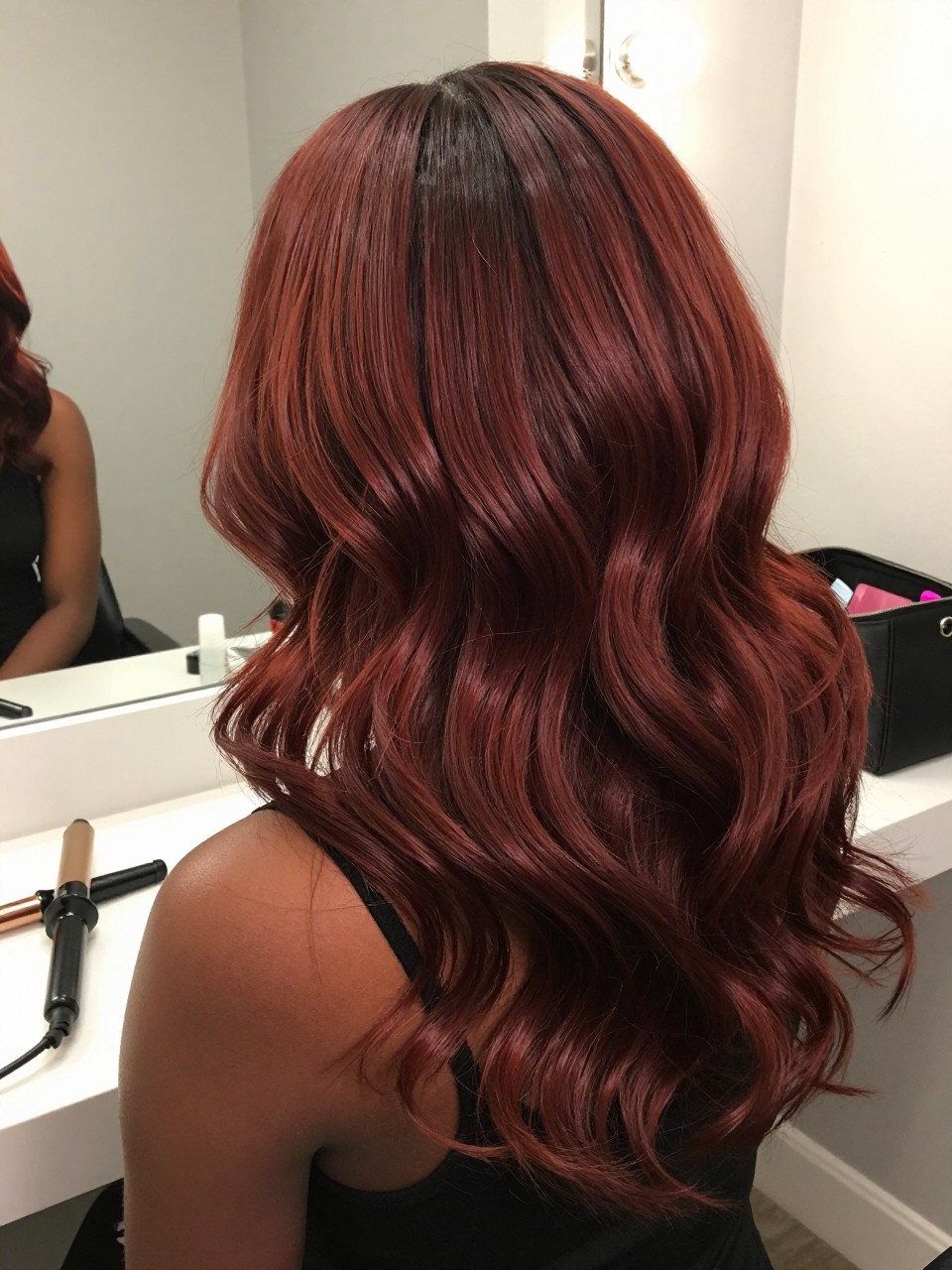 Photo of a black woman wearing rich mahogany root melt hair with deeper roots and warm red-brown lengths in loose brushed waves, back-of-head view to highlight the blended root melt, bathroom vanity mirror setting, soft indoor lighting, with a curling iron on the counter and a makeup bag nearby, latest iPhone photo quality.