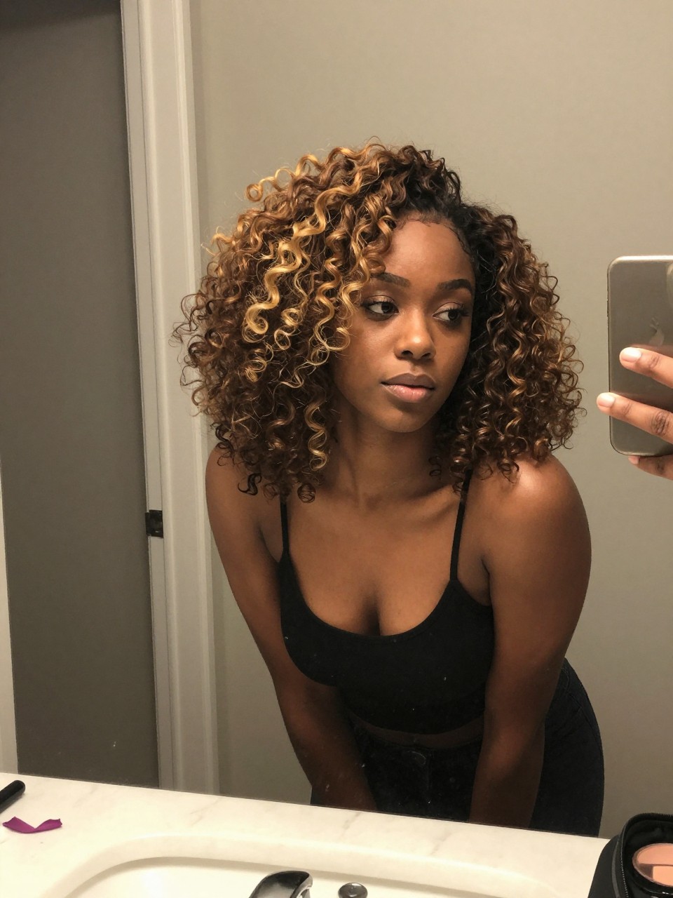 Photo of a Black woman wearing a half-up natural puff with golden highlights concentrated in the top curls, candid mirror selfie, at a bathroom vanity mirror, soft indoor lighting, containing a small lived-in detail such as a ribbon on the counter and a makeup bag nearby, latest iPhone photo quality.
