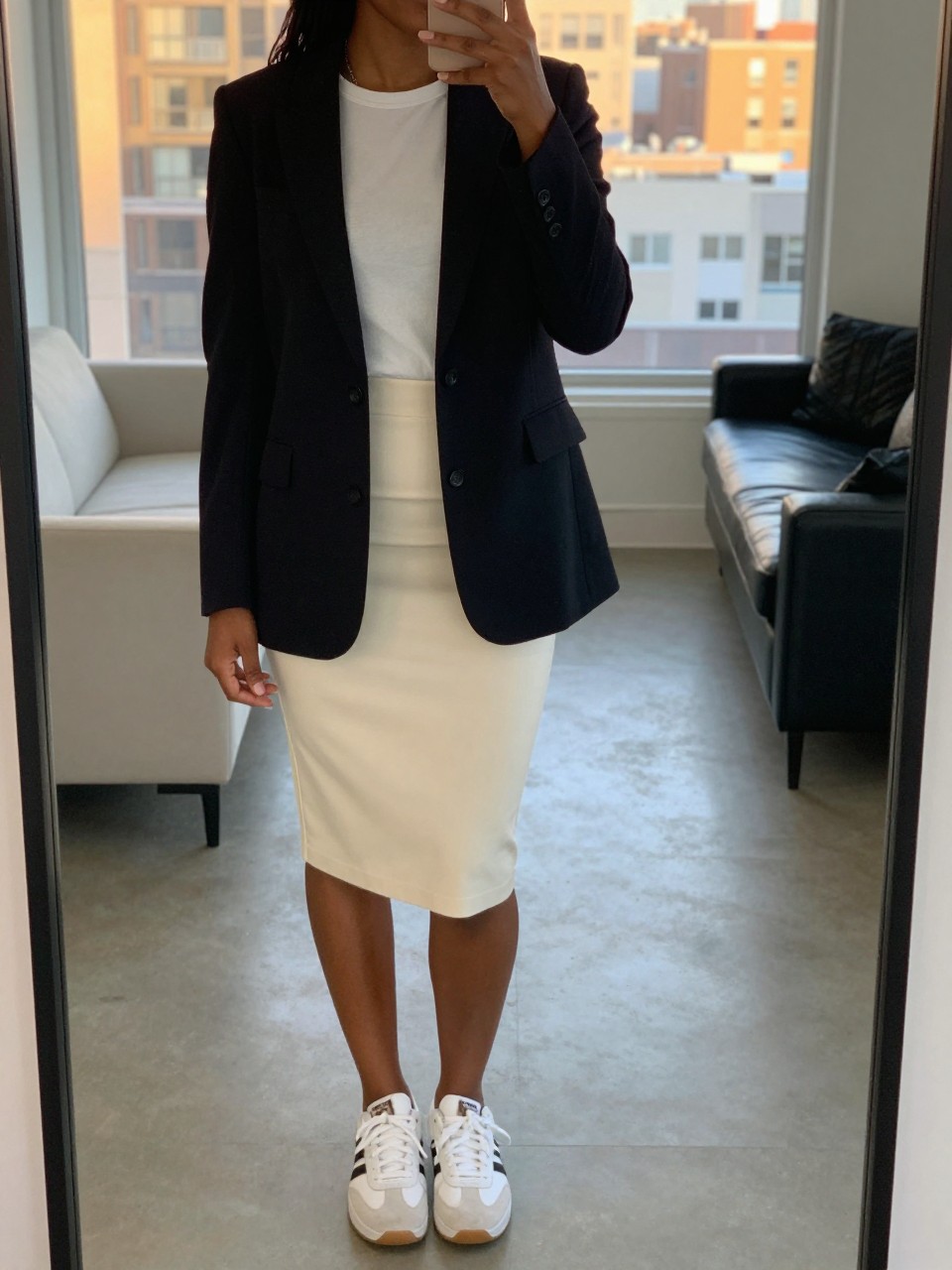 mirror selfie of a black woman wearing a black blazer over a white tee and cream pencil skirt with white sneakers, holding her phone up near her shoulder, cropped at NECK so face doesn't show, in a bright city apartment, evening golden hour, iPhone photo quality.