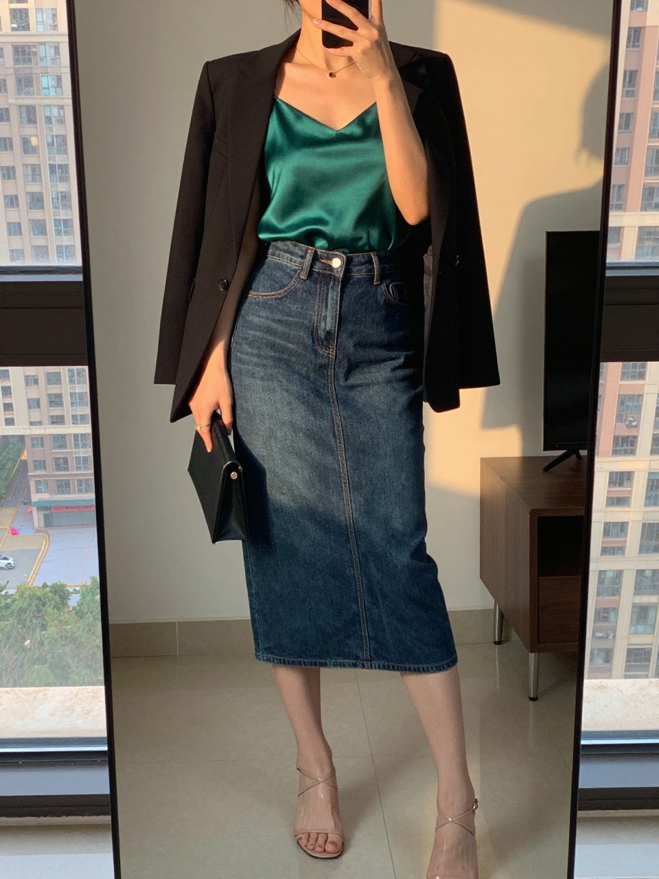 iPhone mirror selfie of a woman wearing an emerald silk camisole tucked into a dark-wash denim midi skirt with a black blazer draped over her shoulders and nude strappy heels, holding a small clutch, standing in a confident pose, cropped at NECK so face doesn't show, in a city apartment, evening golden hour, iPhone photo quality.