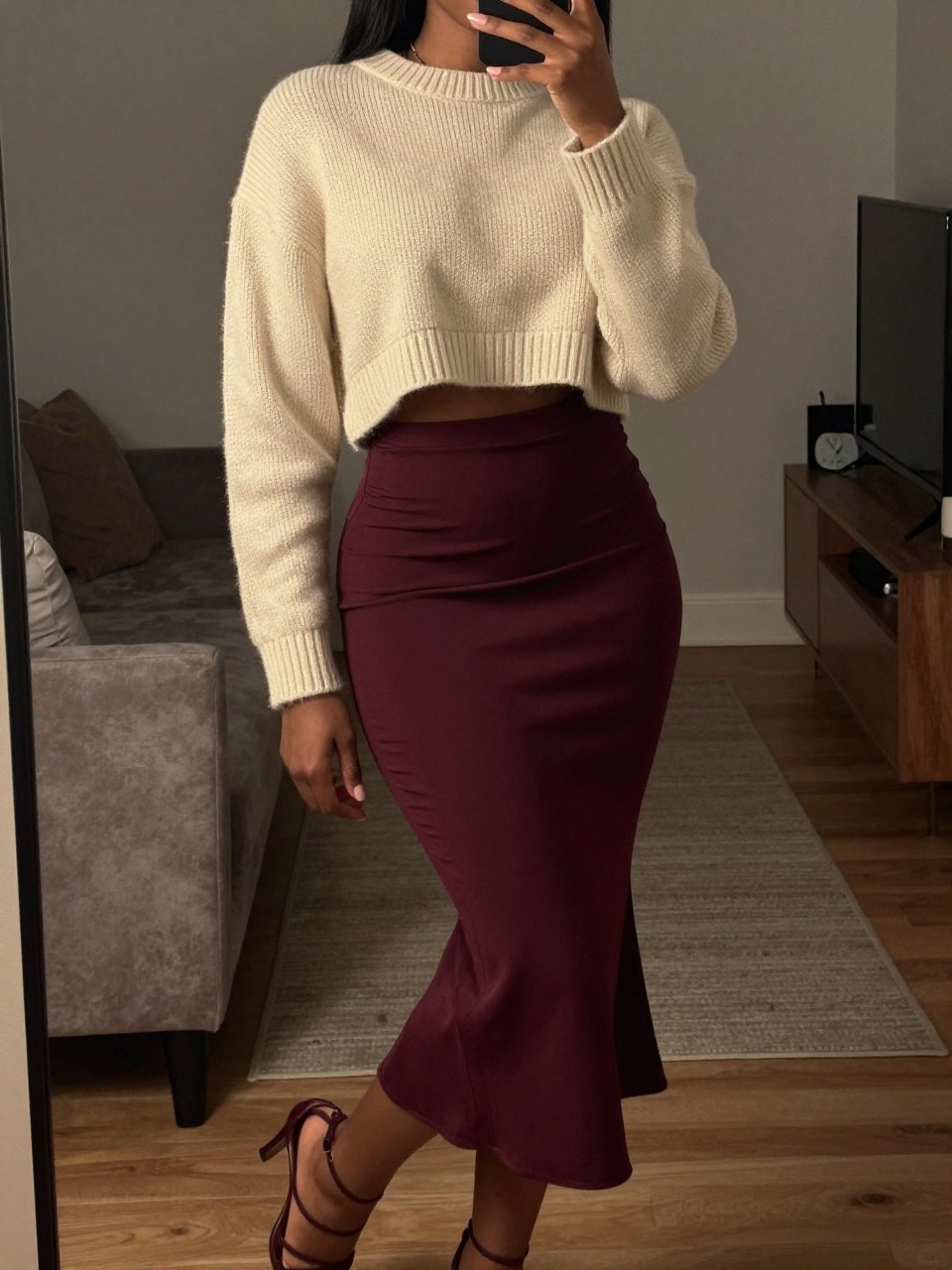 mirror selfie of a black woman in a cream cropped cashmere sweater and a burgundy mermaid skirt with strappy heels, posing with one foot forward, cropped at NECK so face doesn't show, in a city apartment, warm moody lighting, iPhone photo quality.