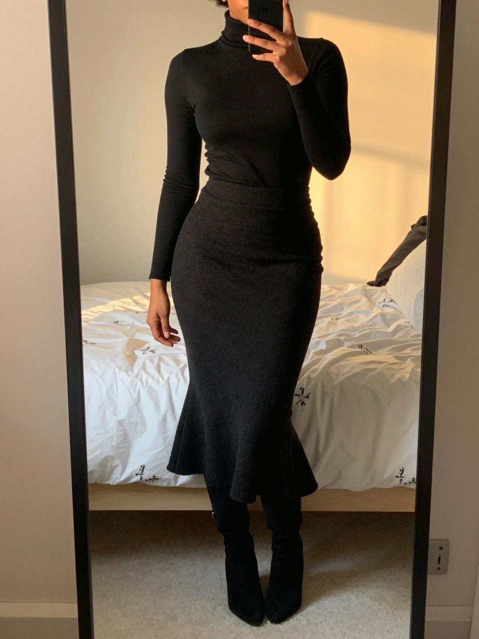 iPhone mirror selfie of a black woman in a black turtleneck and a black wool mermaid skirt with over-the-knee boots, standing with her weight on one leg, cropped at NECK so face doesn’t show, in a feminine-themed bedroom, evening golden hour, iPhone photo quality