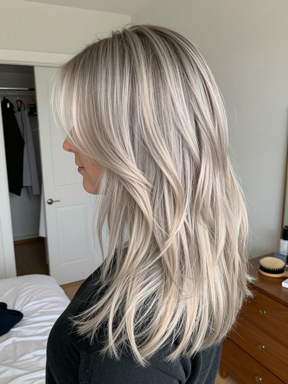 Photo of a woman wearing a layered grey blonde blowout with icy ashy highlights and soft bouncy ends, side profile view, in a sun-drenched bedroom, natural window light, containing a small lived-in detail such as a slightly open closet behind and a round brush on the dresser, latest iPhone photo quality.