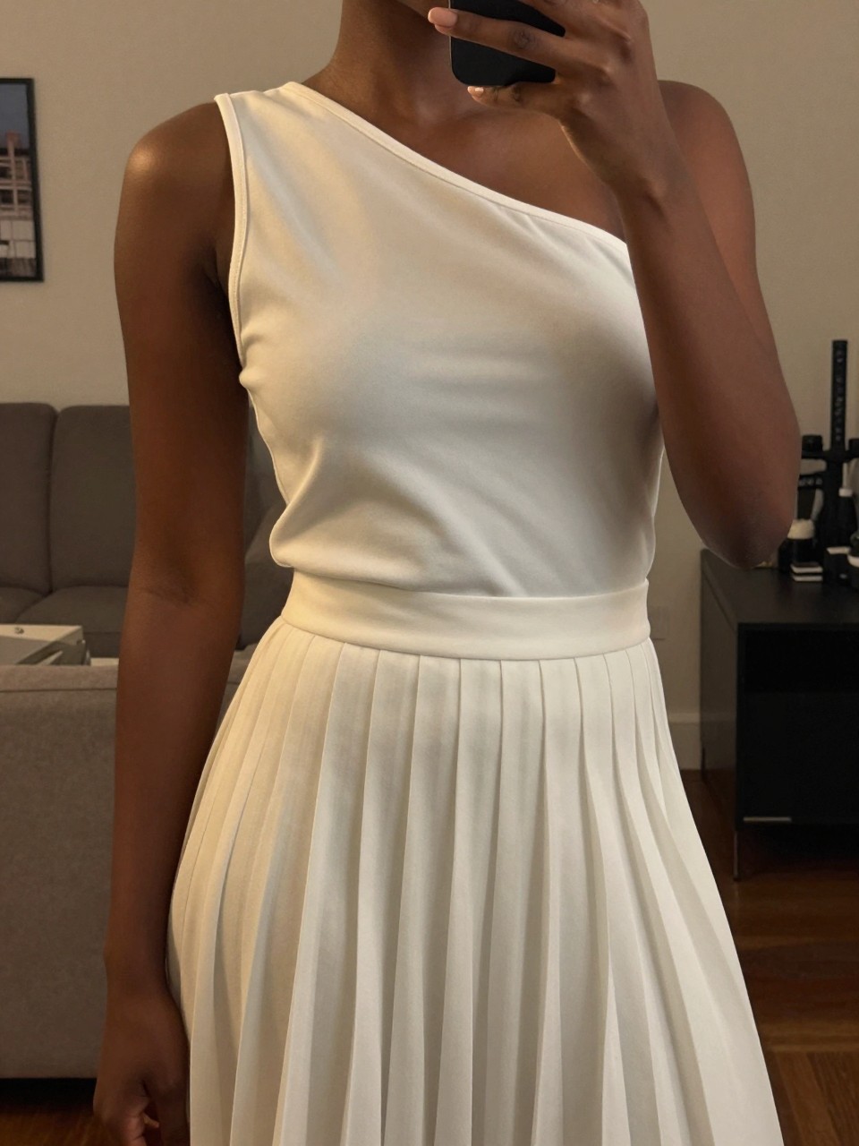 iPhone mirror selfie of a black woman in an asymmetrical top and white pleated skirt, showing the shoulder detail, cropped at NECK so face doesn't show, in a city apartment, warm moody lighting, iPhone photo quality.