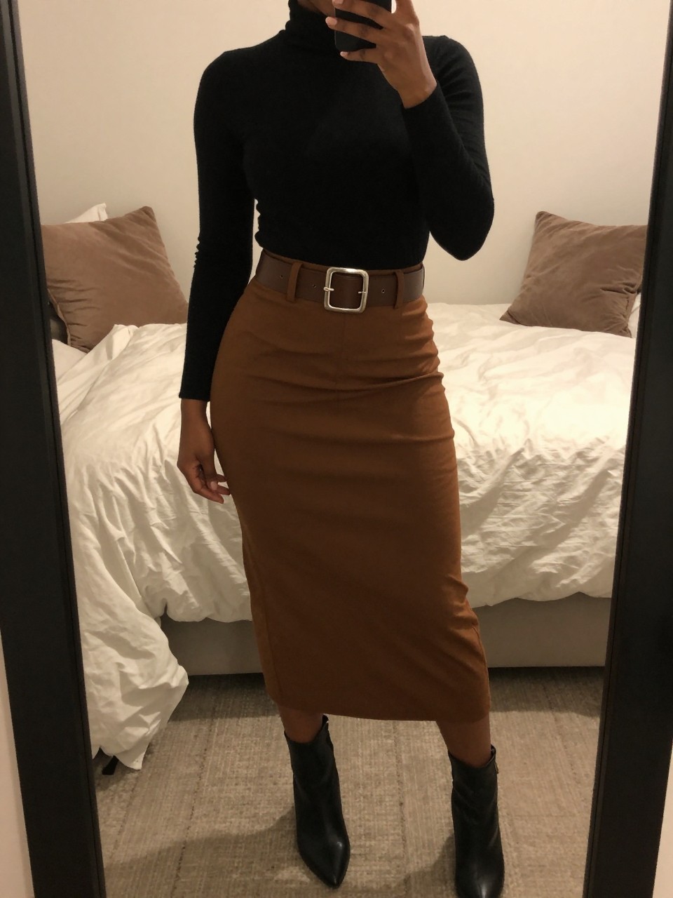 mirror selfie of a black woman wearing a black turtleneck tucked into a brown midi skirt with a wide silver-buckle belt and black ankle boots, one hand resting on the belt, cropped at NECK so face doesn't show, in a feminine-themed bedroom, warm moody lighting, iPhone photo quality.