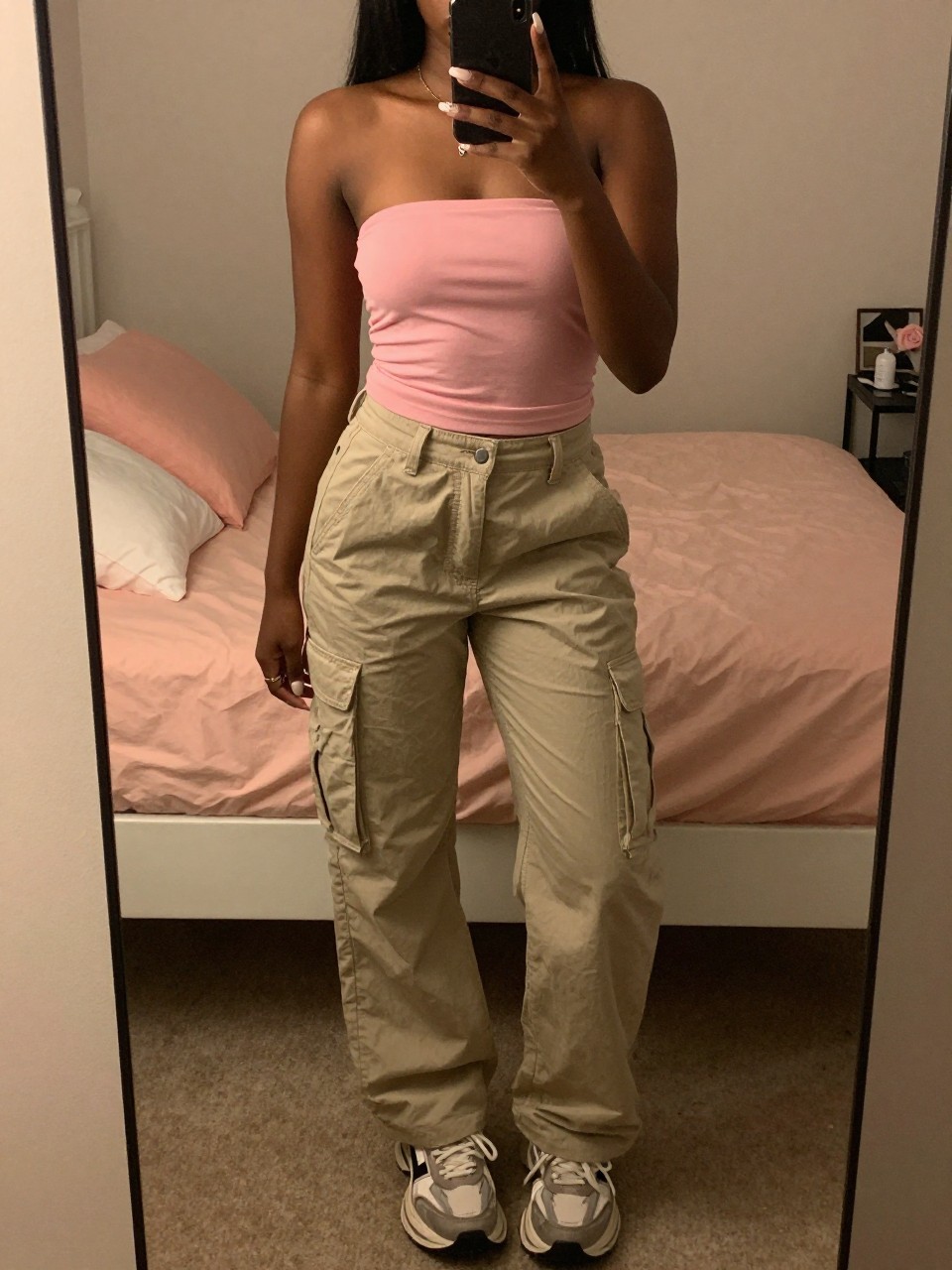 iPhone mirror selfie of a black woman in a pink tube top with cargo pants and chunky sneakers, standing with phone at chest, cropped at NECK so face doesn't show, in a feminine-themed bedroom, warm moody lighting, iPhone photo quality.