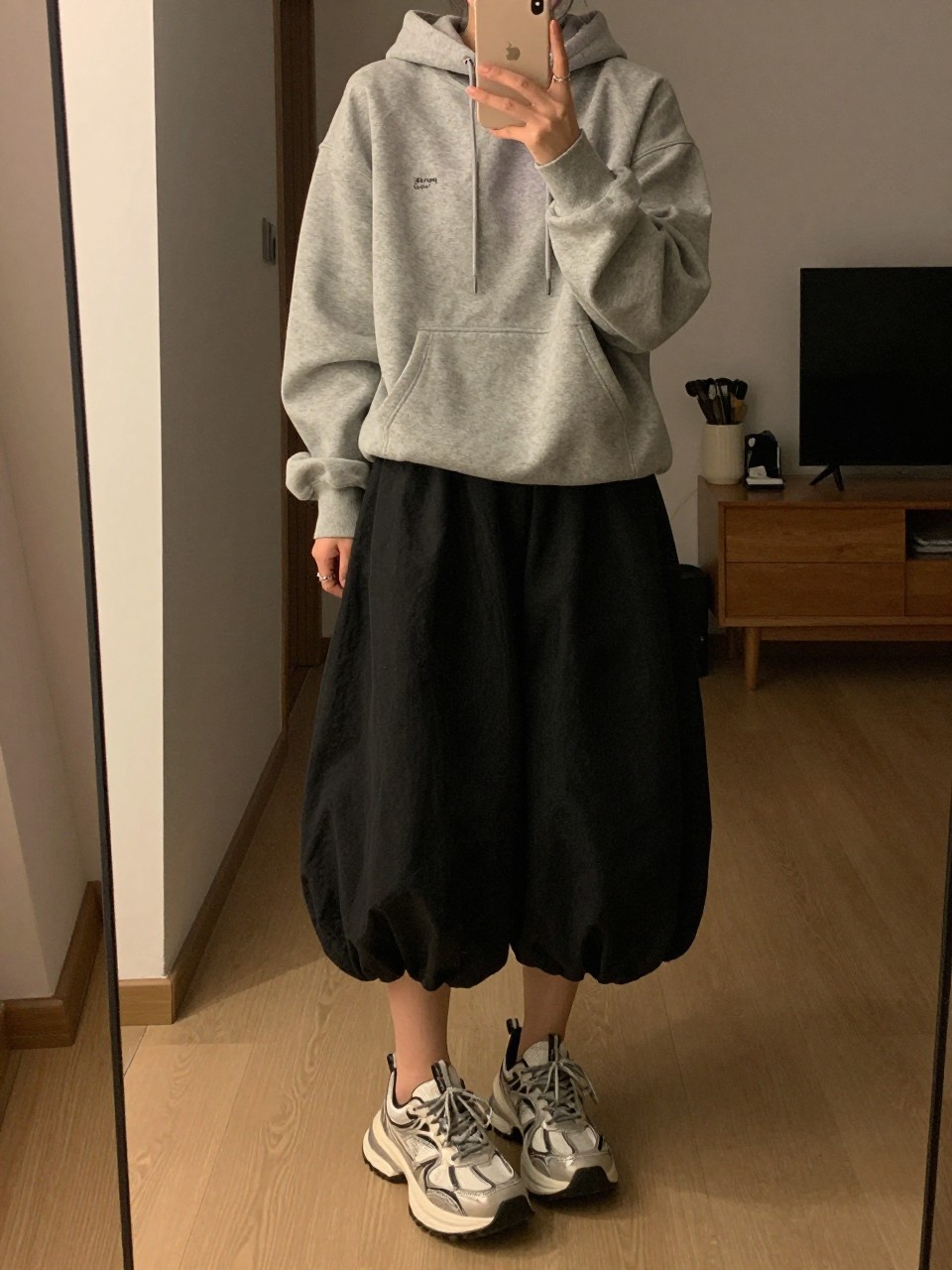iPhone mirror selfie of a woman in a grey hoodie and a black balloon skirt with chunky dad sneakers, phone in hand, cropped at NECK so face doesn't show, in a city apartment, warm moody lighting, iPhone photo quality.
