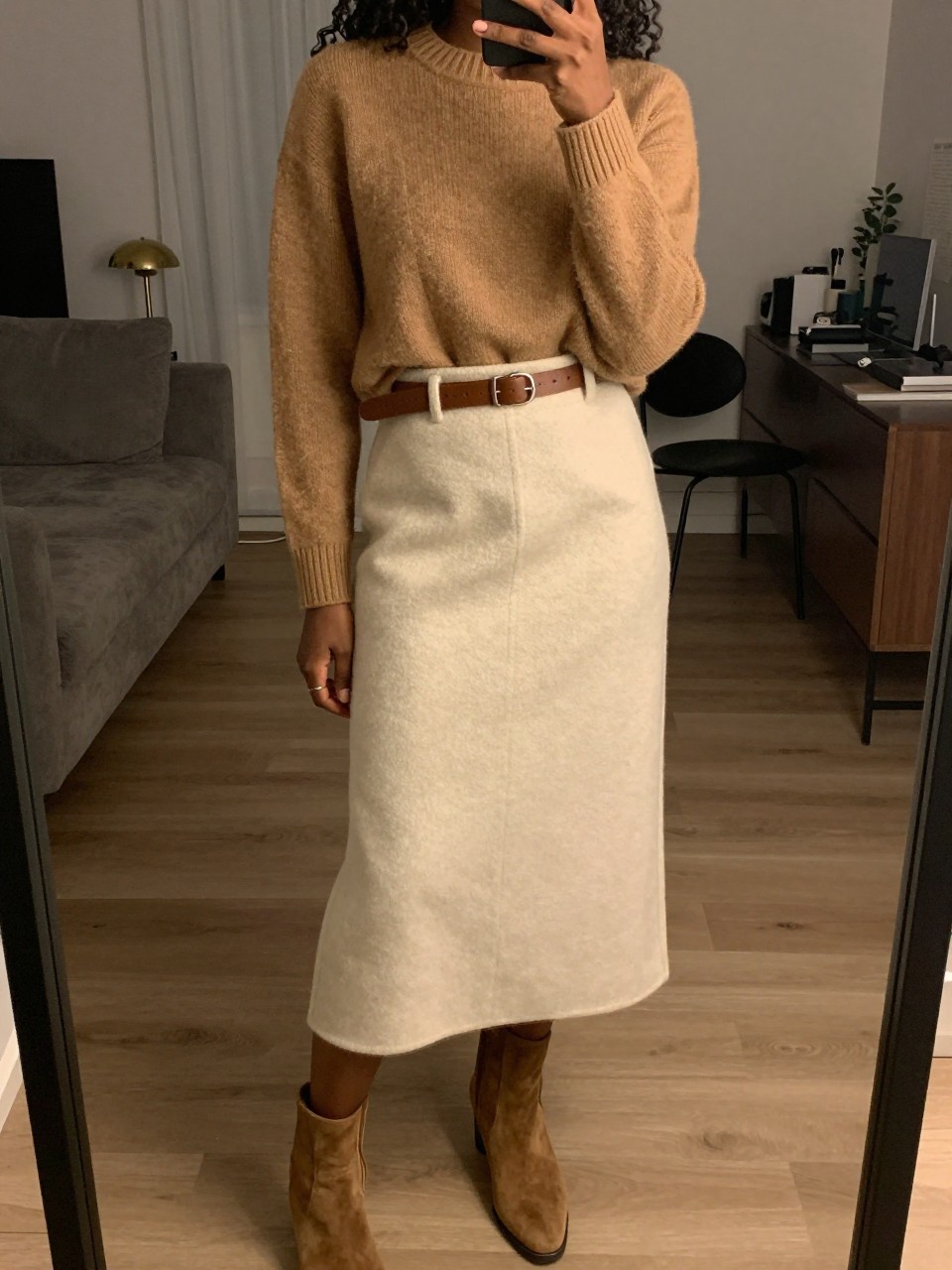 mirror selfie of a black woman wearing a camel crewneck sweater tucked into a soft cream wool midi skirt with a brown leather belt and matching suede ankle boots, one hand resting on the belt, cropped at NECK so face doesn't show, in a city apartment, warm moody lighting, iPhone photo quality.
