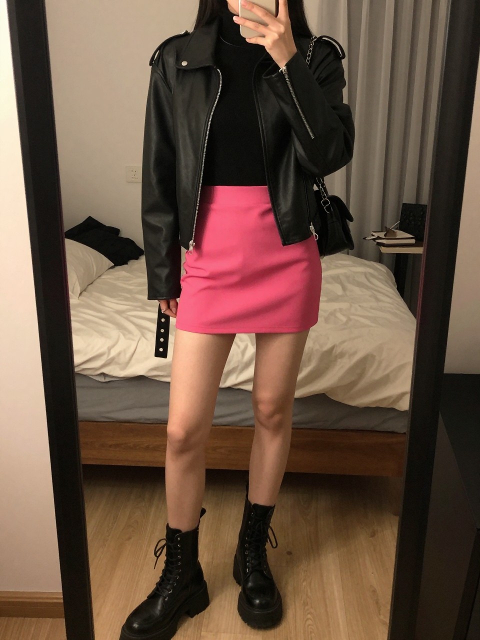 iPhone mirror selfie of a woman wearing a black turtleneck tucked into a hot pink mini skirt with black chunky combat boots and a black leather jacket worn open, a silver chain visible, standing in an edgy pose with hands in pockets, cropped at NECK so face doesn't show, in a feminine-themed bedroom, warm moody lighting, iPhone photo quality.
