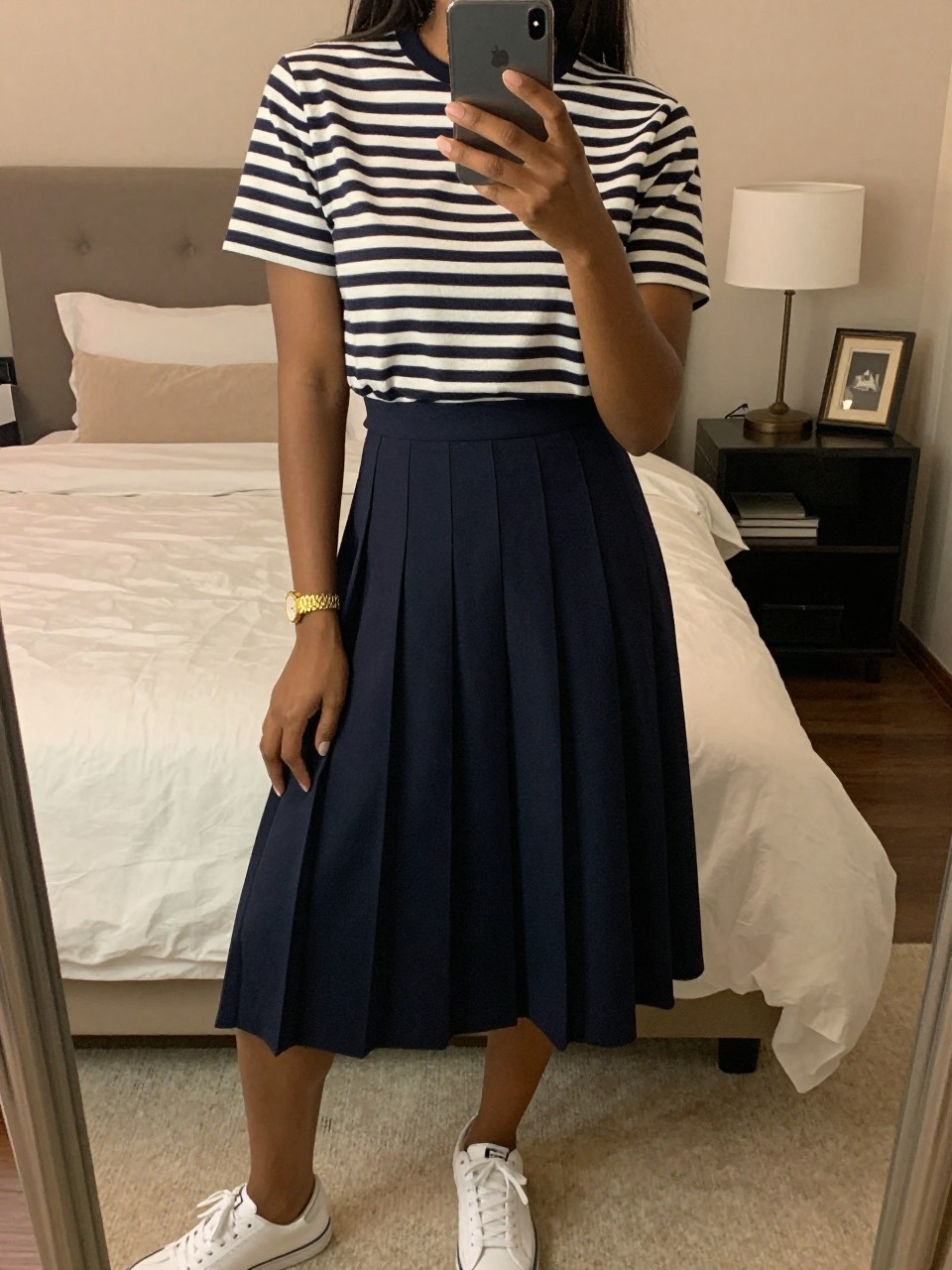iPhone mirror selfie of a black woman wearing a navy striped Breton top tucked into a navy pleated midi skirt with a gold watch and white sneakers, holding her phone low by her thigh, cropped at NECK so face doesn't show, in a feminine-themed bedroom, warm moody lighting, iPhone photo quality.