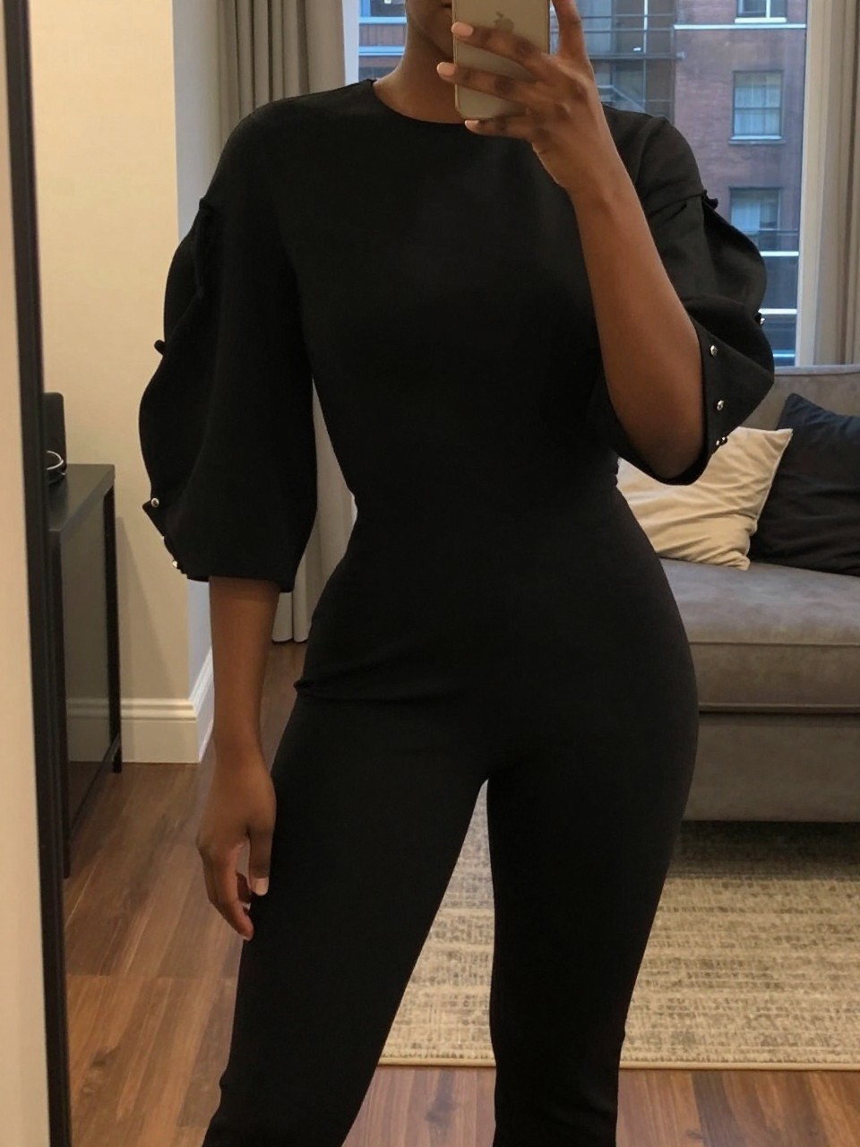 mirror selfie of a black woman in a black jumpsuit with statement sleeves, showcasing the sleeve detail, cropped at NECK so face doesn't show, in a city apartment, warm moody lighting, iPhone photo quality.