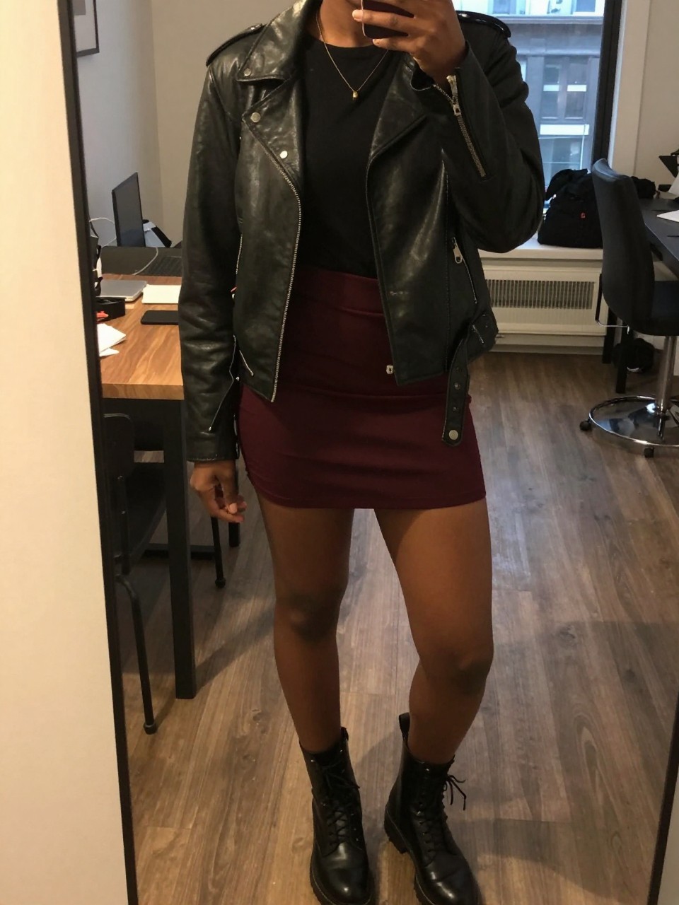 mirror selfie of a black woman wearing a black leather jacket over a band tee with a deep burgundy mini skirt and black combat boots, one hand in her jacket pocket, cropped at NECK so face doesn't show, in a city apartment, warm moody lighting, iPhone photo quality.